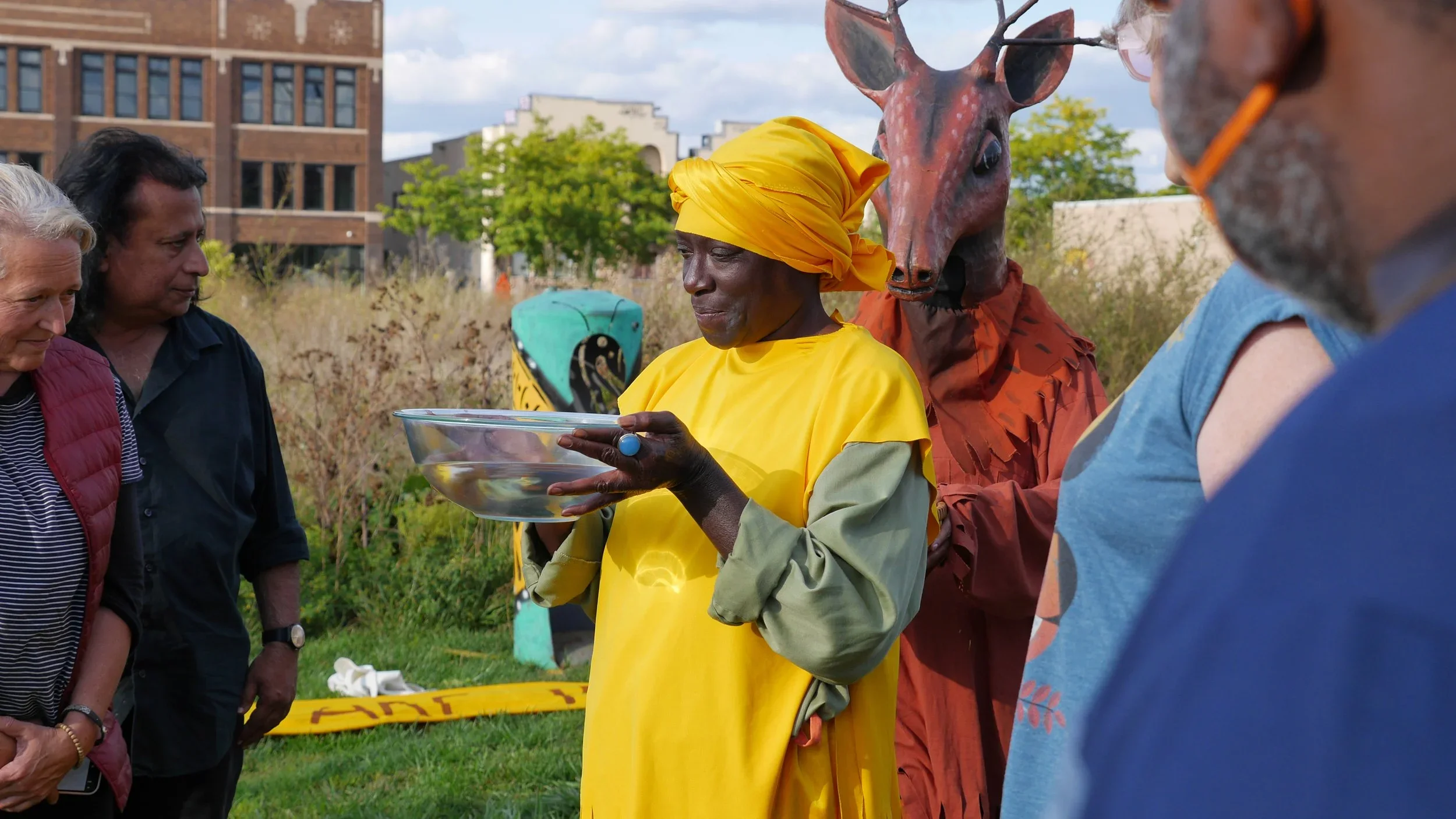 A group of people gathered outdoors, with some dressed in colorful costumes, including a person in a yellow outfit with a matching headscarf and a person in a giraffe mascot costume. The scene appears to be a cultural or festive event in a park with trees and buildings in the background.