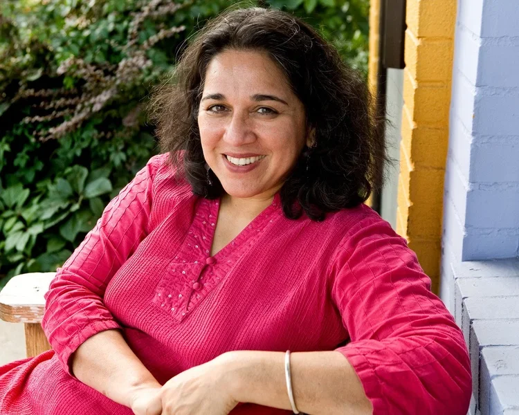Woman with dark curly hair wearing a pink top, sitting outdoors on a porch with a green hedge in the background, smiling at the camera.