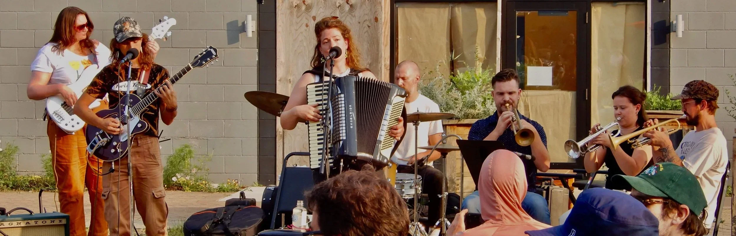 Group of musicians performing outdoors, including a woman playing an accordion and singing into a microphone, a guitarist, trumpet players, and a drummer. Audience members are visible in the foreground.