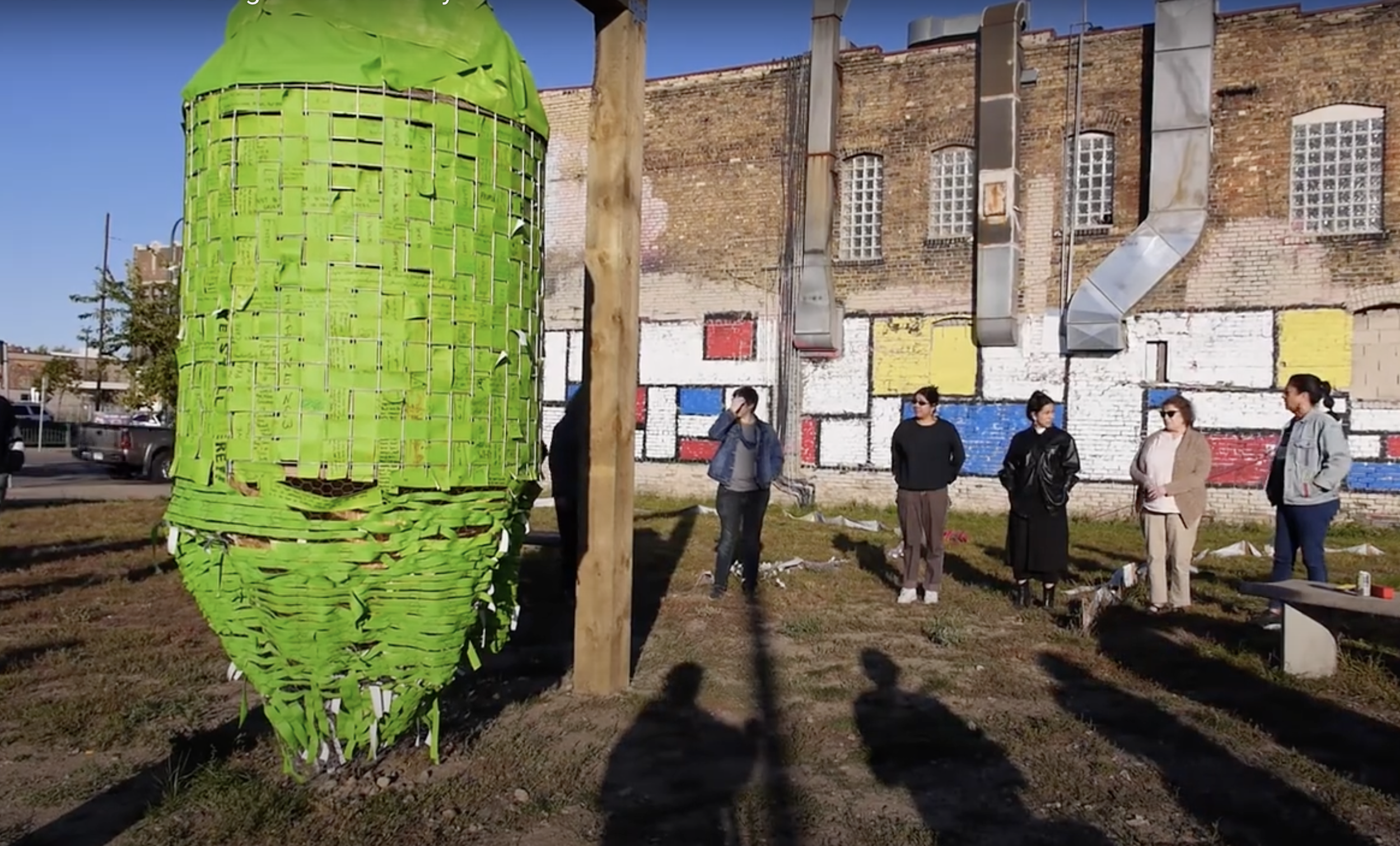A group of six people standing outdoors in front of a brick wall with a colorful mural, with a large green mask-shaped sculpture made of paper or cardboard strips in the foreground to the left.