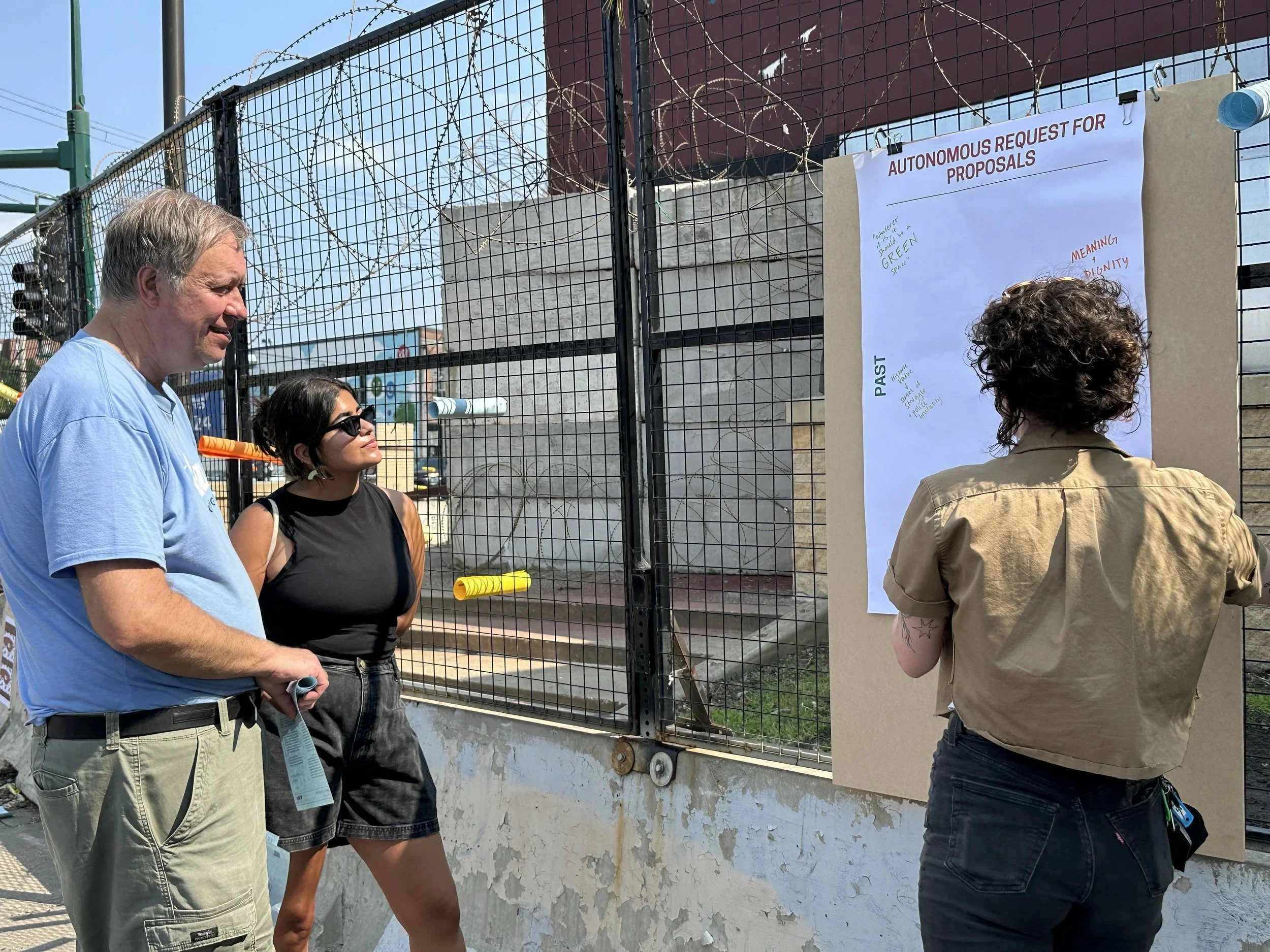 Three people standing outdoors near a fence with barbed wire, looking at a large proposal poster on a board. The poster is titled "Autonomous Request for Proposals" and has handwritten notes. The setting appears to be an outdoor event or protest.