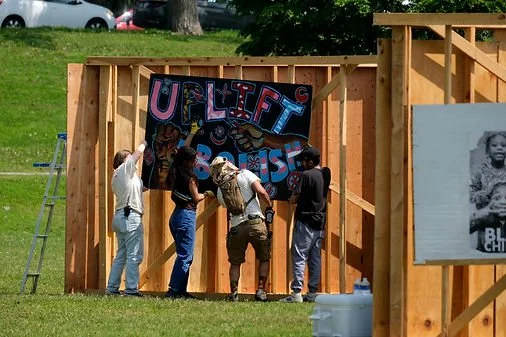 Four people working on a construction site installing a large colorful banner that says 'Uplift Black' on a wooden frame.