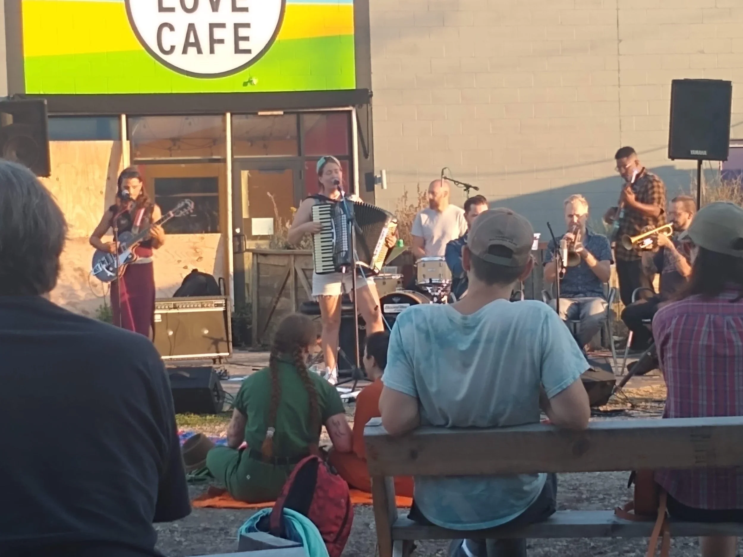 A live outdoor band performance at Love Cafe with multiple musicians playing instruments, including guitar, accordion, drums, trumpet, and saxophone, during the daytime with an audience seated on benches and on the ground.