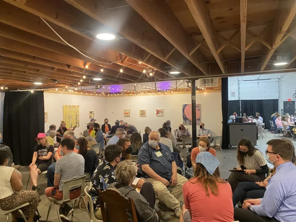 People sitting and chatting in an indoor event space with art on the walls, string lights, and a black curtain dividing the room.