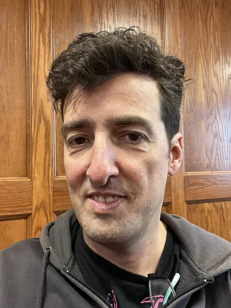 A close-up of a man with dark, curly hair, wearing a black shirt and a gray hoodie, smiling softly in front of a wooden paneled background.