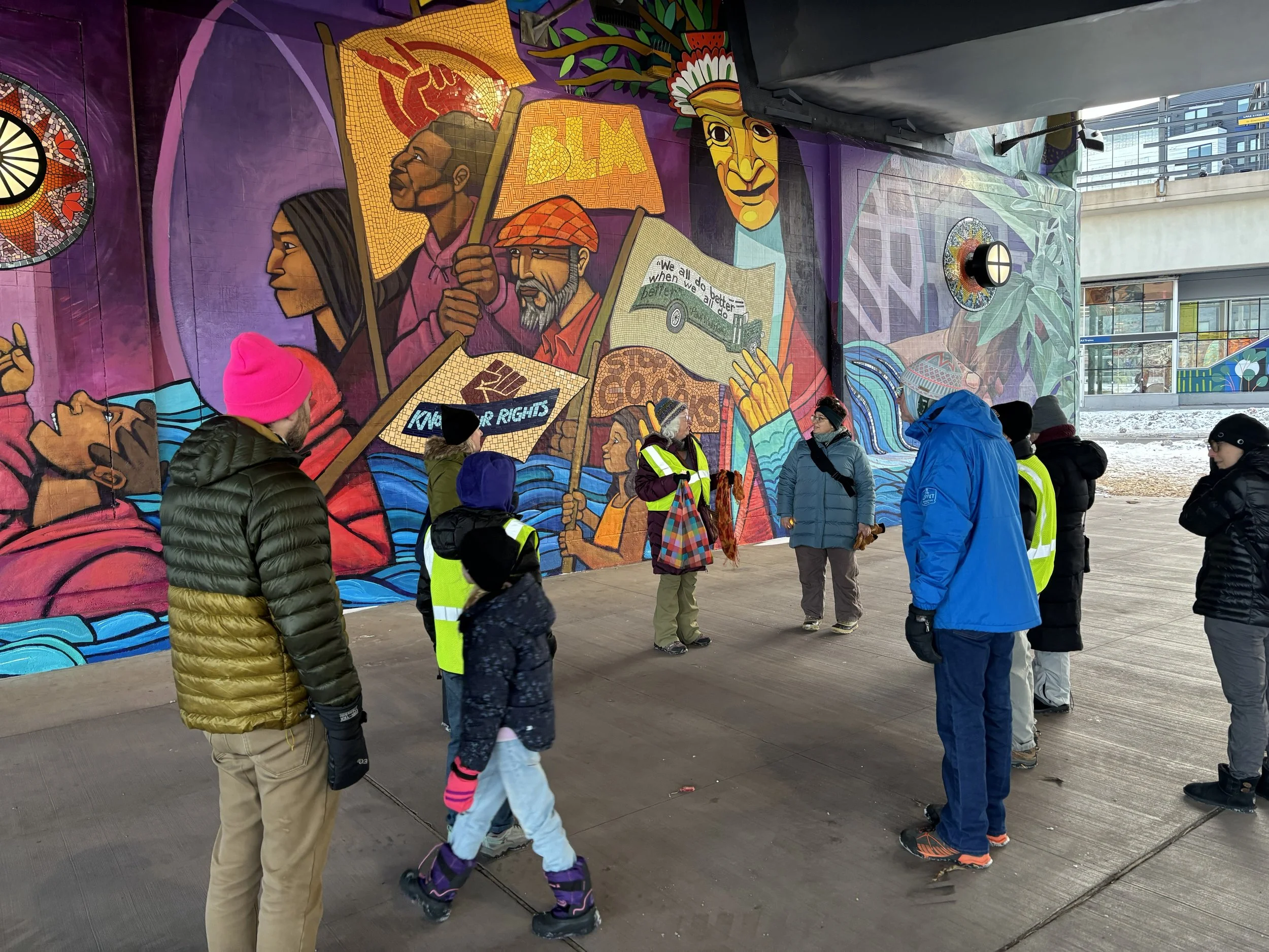A group of people standing in front of a colorful mural depicting people holding signs and banners. The scene appears to be a protest or rally, with some people wearing reflective vests and winter clothing.