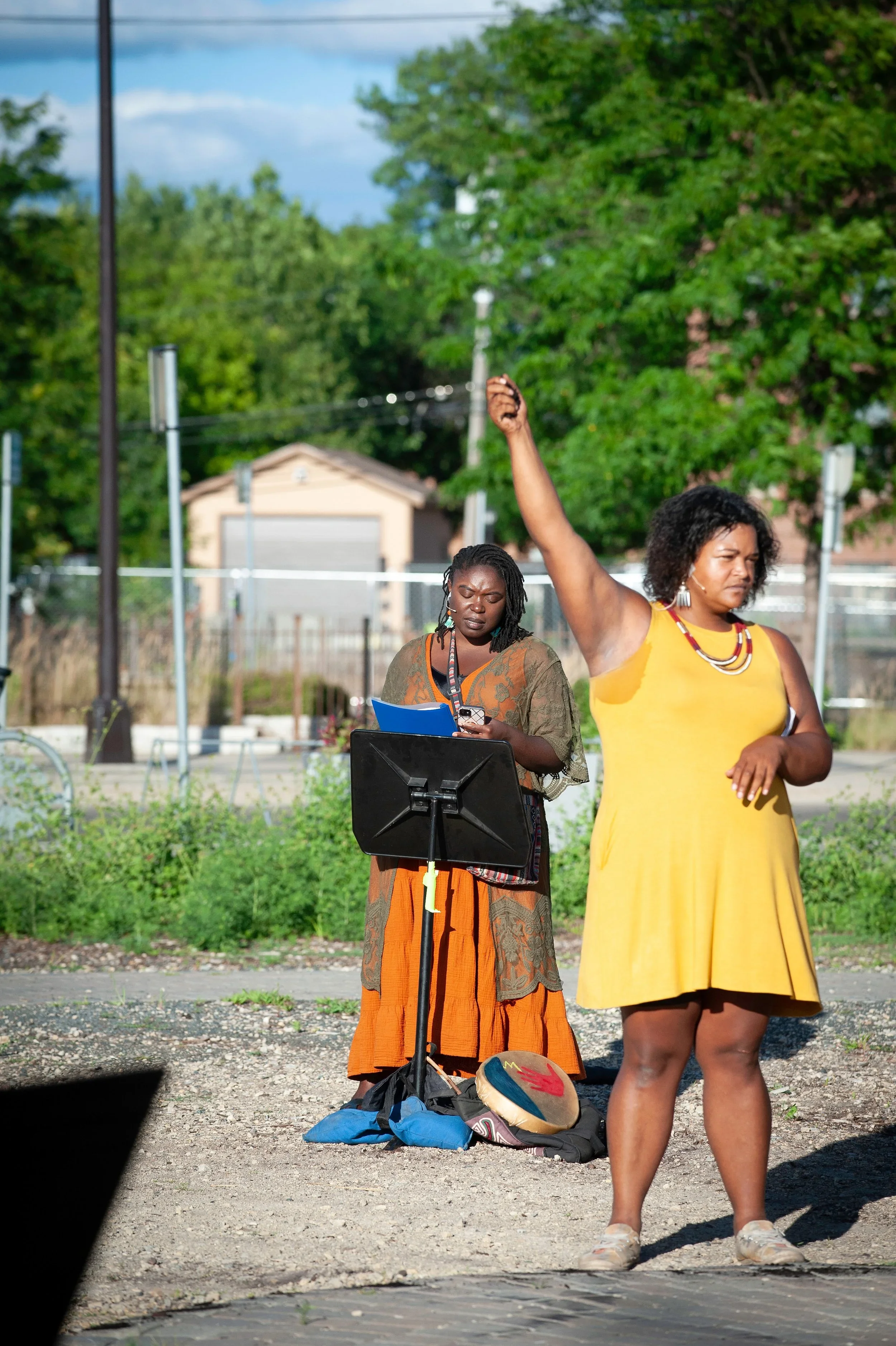 Two women stand outdoors; one is reading sheet music and the other is dancing with her arm raised.