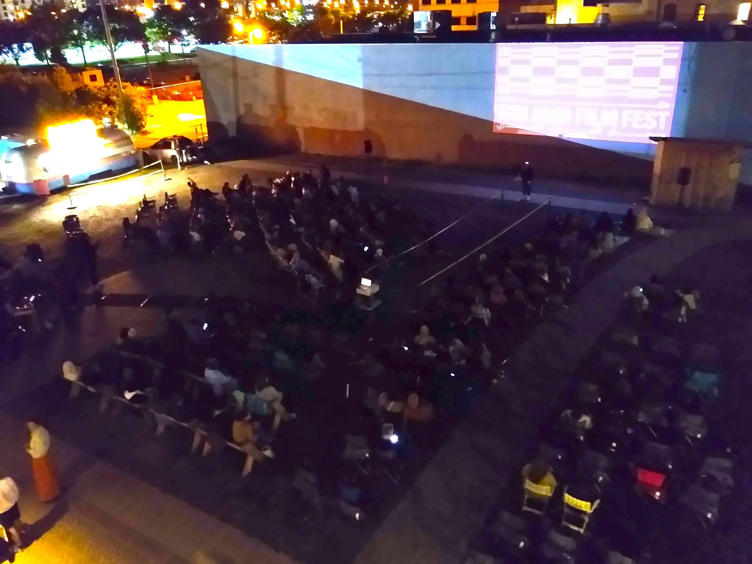 An outdoor movie screening at night with an audience seated on benches and chairs, watching a large screen projected onto a wall.