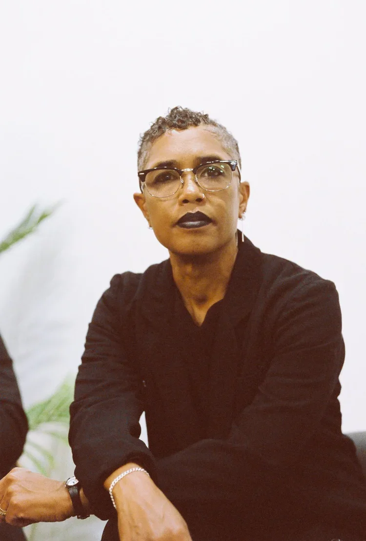 A person with short, curly gray hair wearing glasses with a black frame, dark lipstick, a black shirt, and a bracelet on their wrist, sitting against a plain white background.