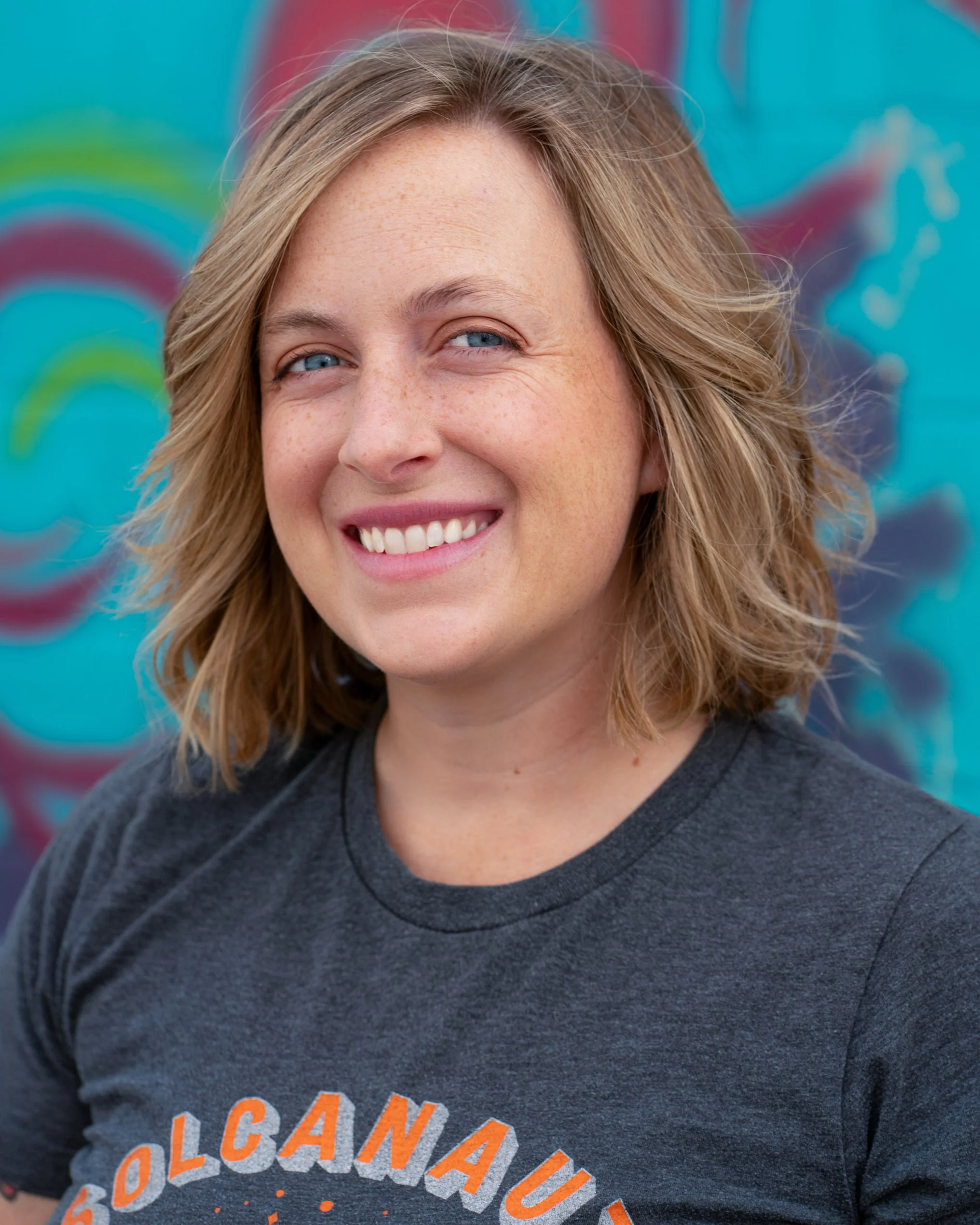 Smiling woman with shoulder-length wavy blonde hair wearing a dark gray T-shirt with orange text, standing in front of a colorful painted mural.