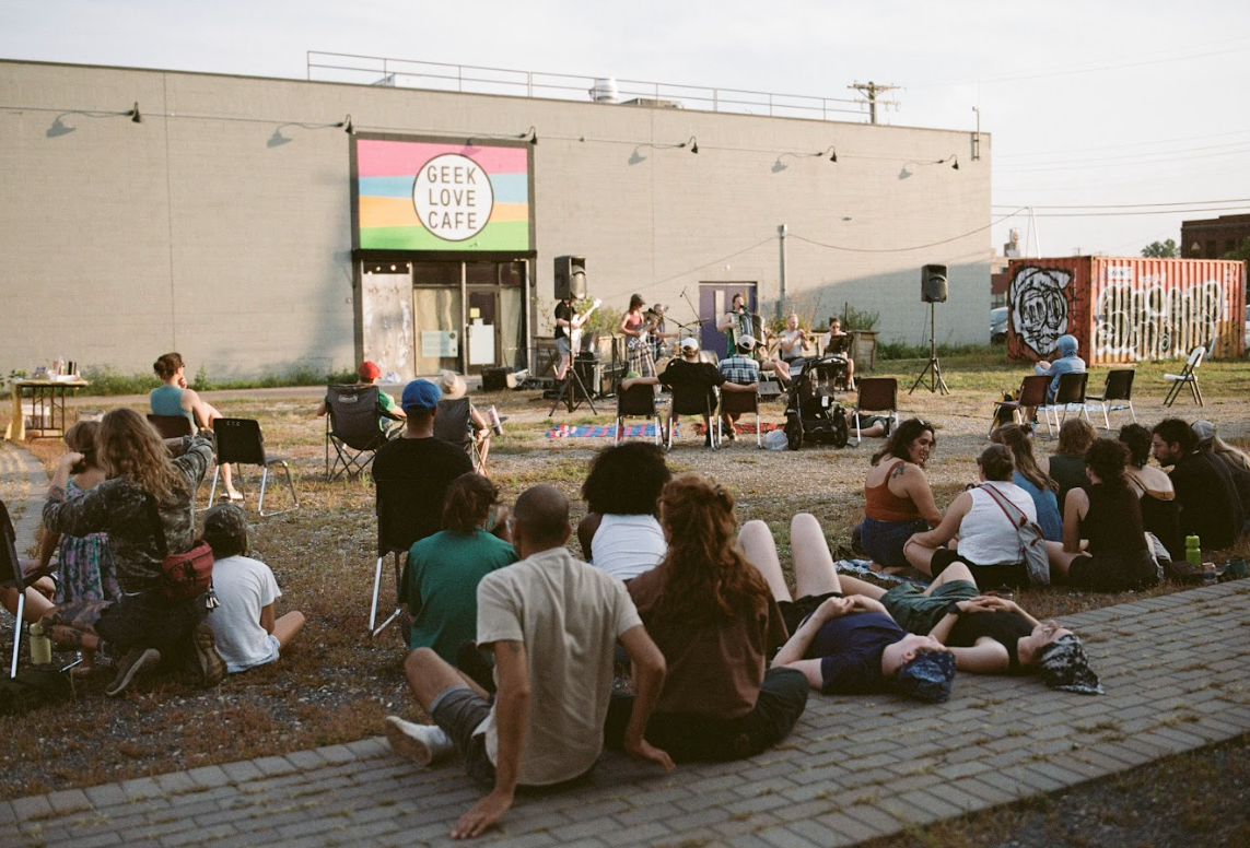Outdoor concert at night with a band playing on a small stage outside a building with a sign that reads 'Geek Love Cafe'. People are sitting on chairs and on the ground, watching the performance.