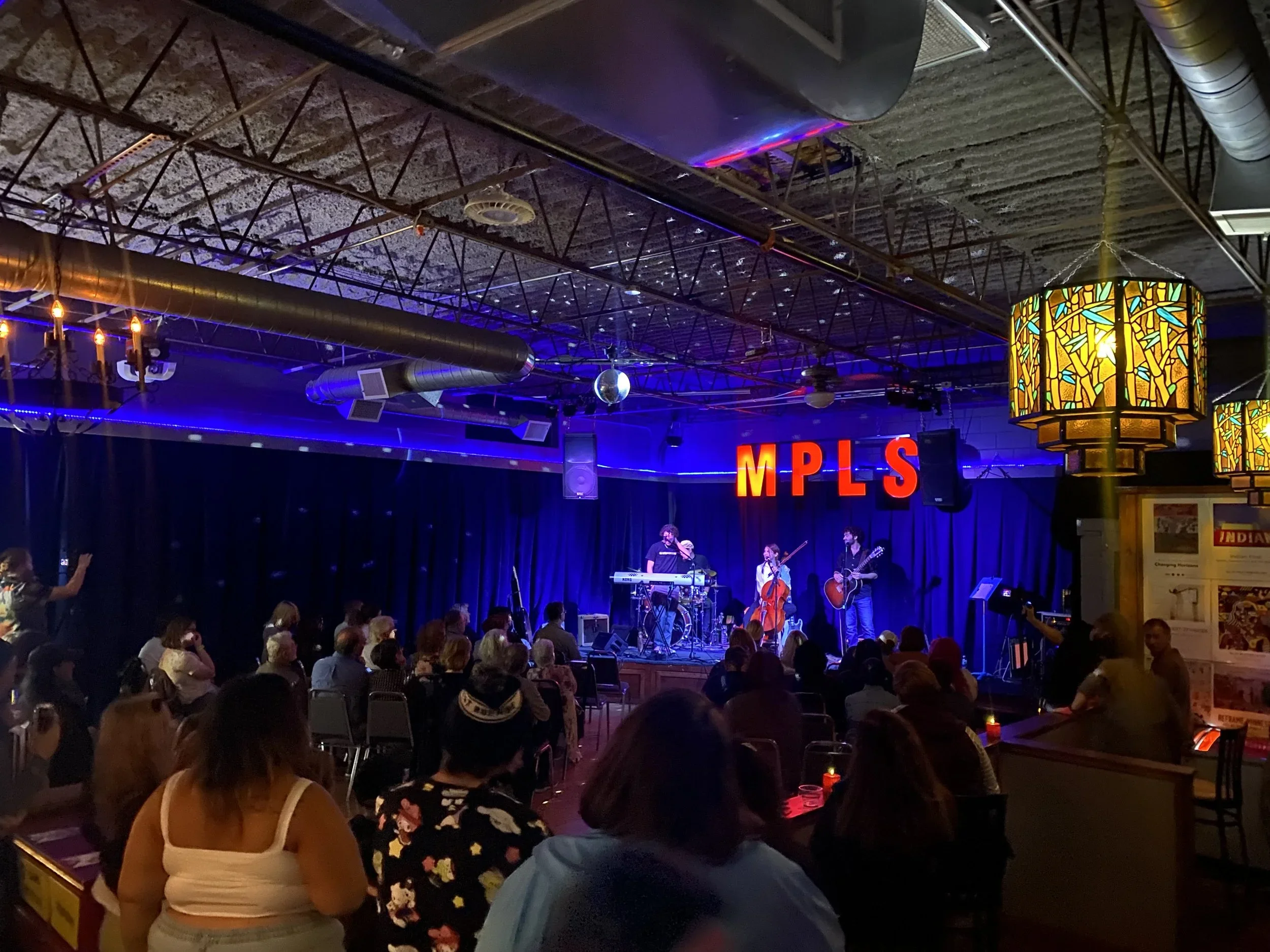Live music performance on stage with three musicians, one playing keyboard, one singing with a cello, and one playing guitar, in a dimly lit venue with blue curtains and red letters spelling 'MPLS' behind them. Audience seated and standing, watching the performance.