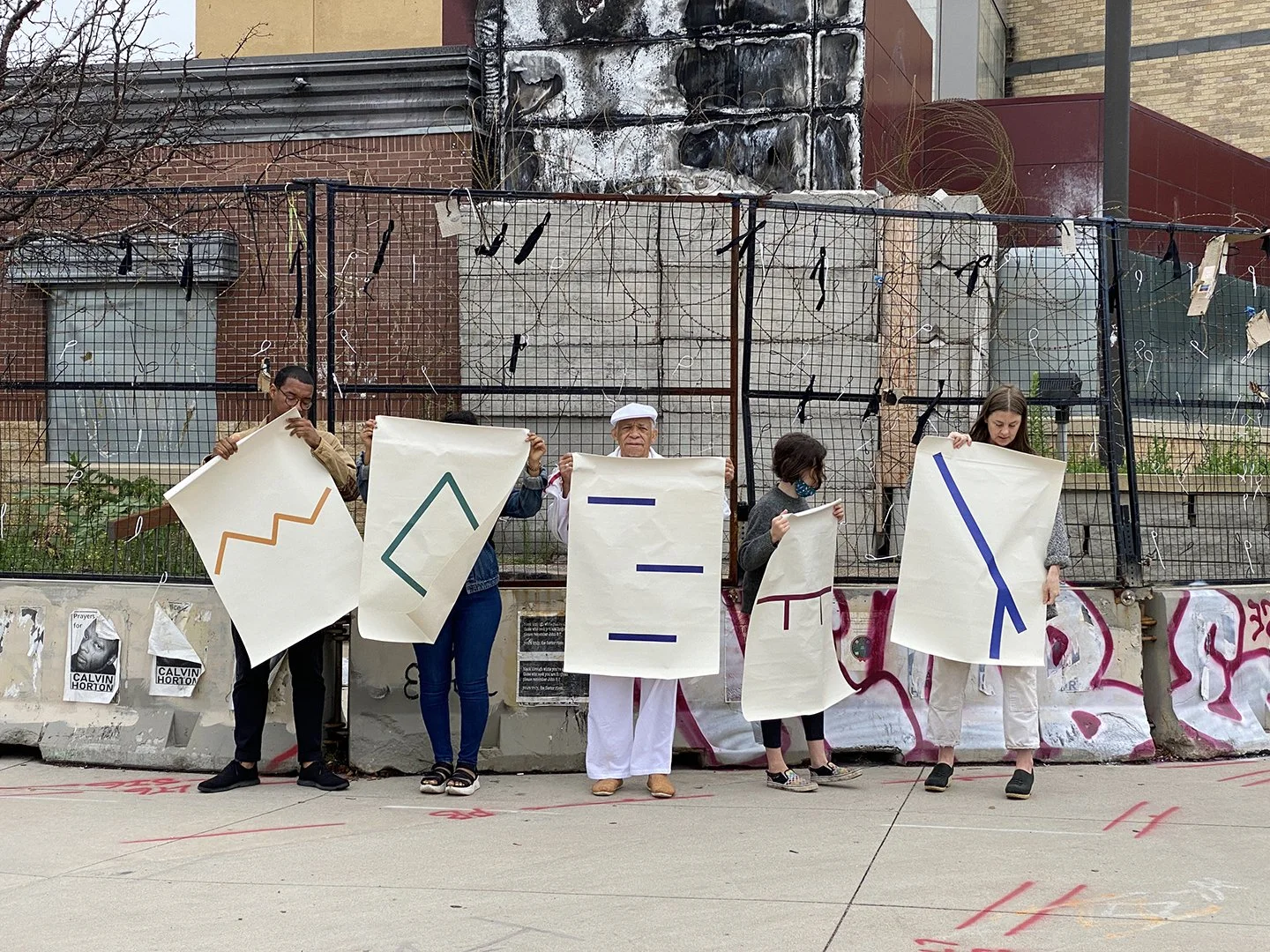 A group of five people standing outdoors, holding large signs with abstract geometric symbols. The background contains a graffiti-covered wall, a metal fence, and urban buildings.
