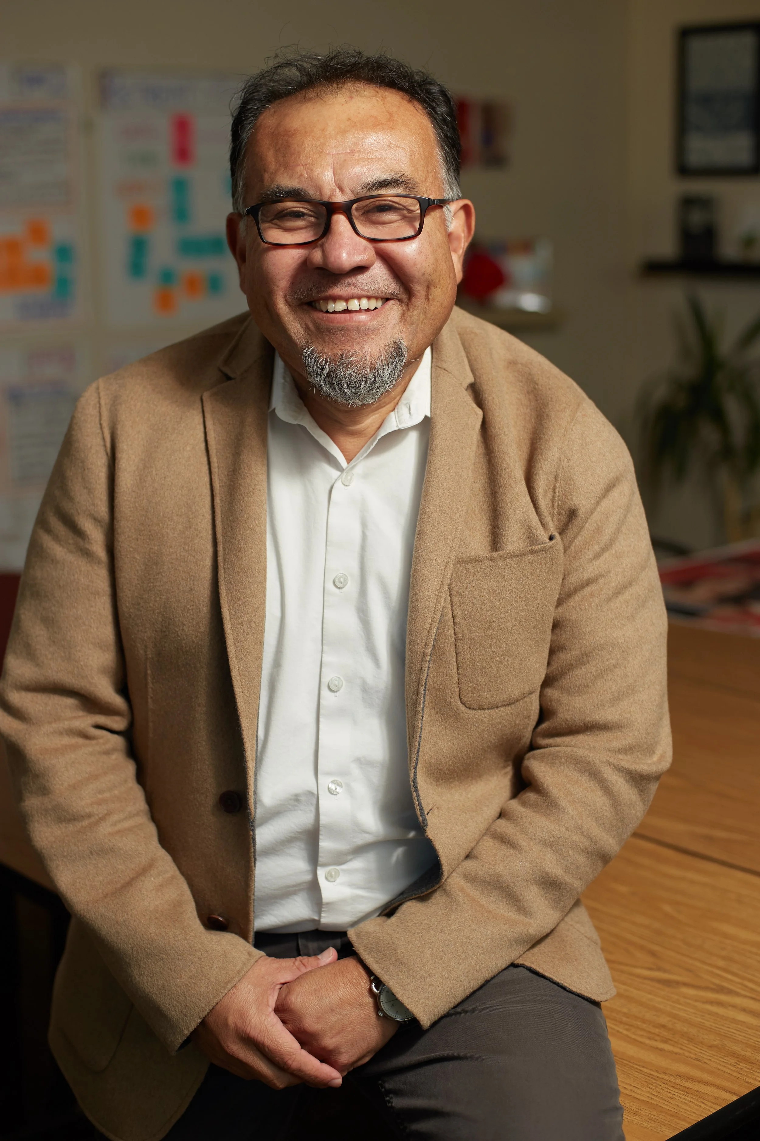 A middle-aged man with glasses, a beard, and a goatee smiling while sitting at a wooden table in an office. He is wearing a white shirt, brown blazer, and a watch. The background features colorful posters and a plant.
