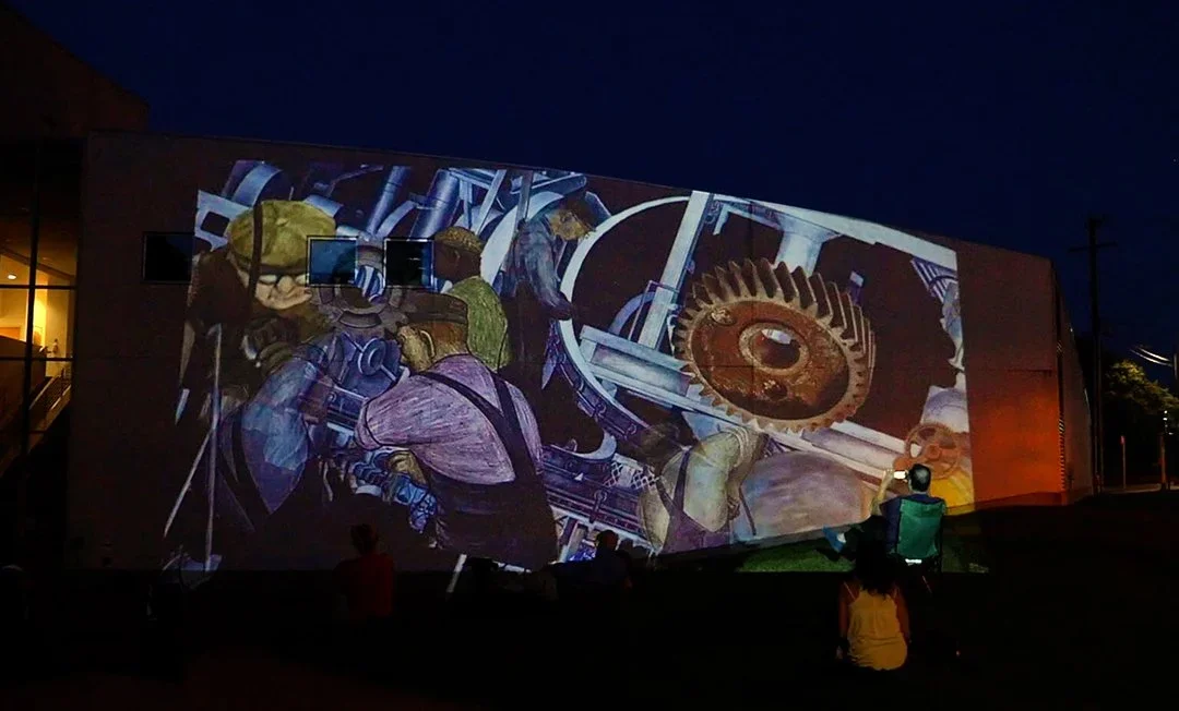 Nighttime outdoor projection of industrial workers and machinery on a building, with people watching.