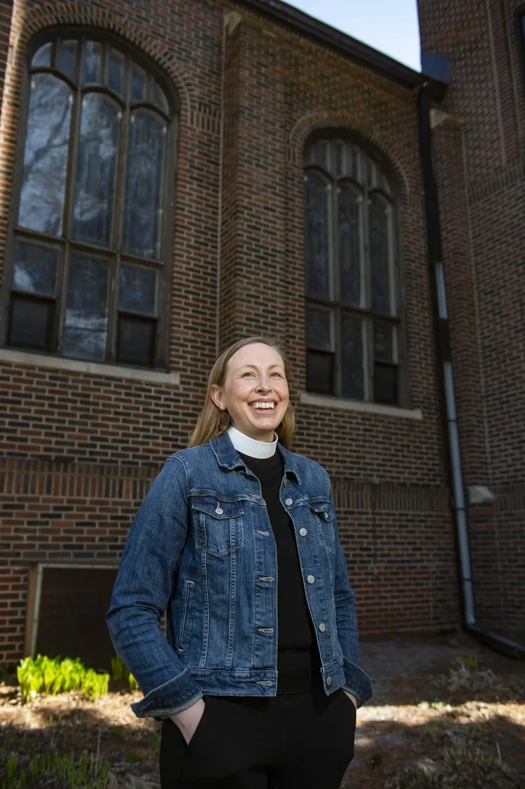 A woman standing outside in front of a brick building with large arched windows, smiling, wearing a denim jacket over a black shirt with a white collar, with sunlight and greenery visible.
