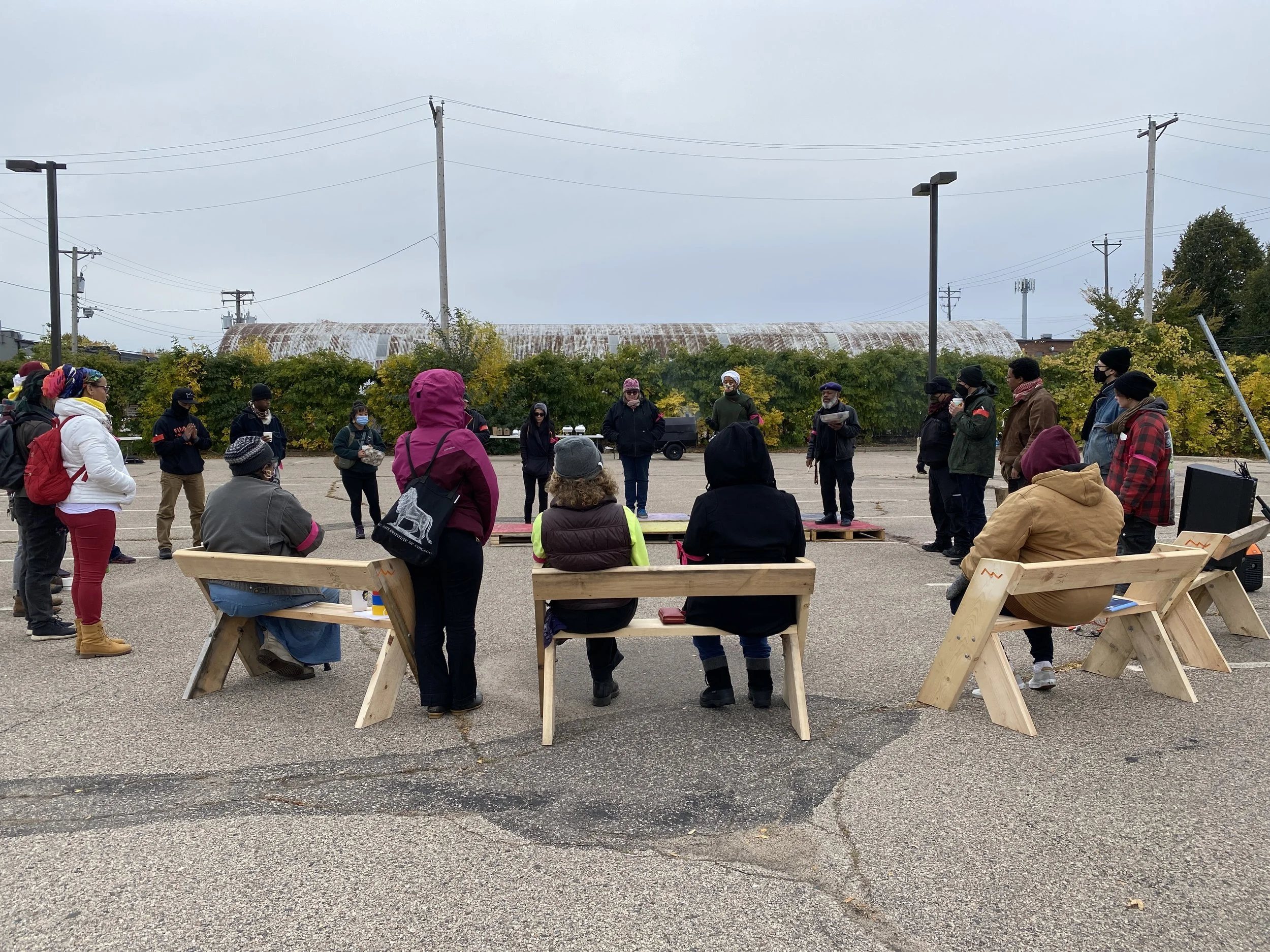 Group of people gathered outdoors in a parking lot, some sitting on wooden benches and others standing, attending a meeting or event under overcast skies, with power lines and a rusted industrial building in the background.