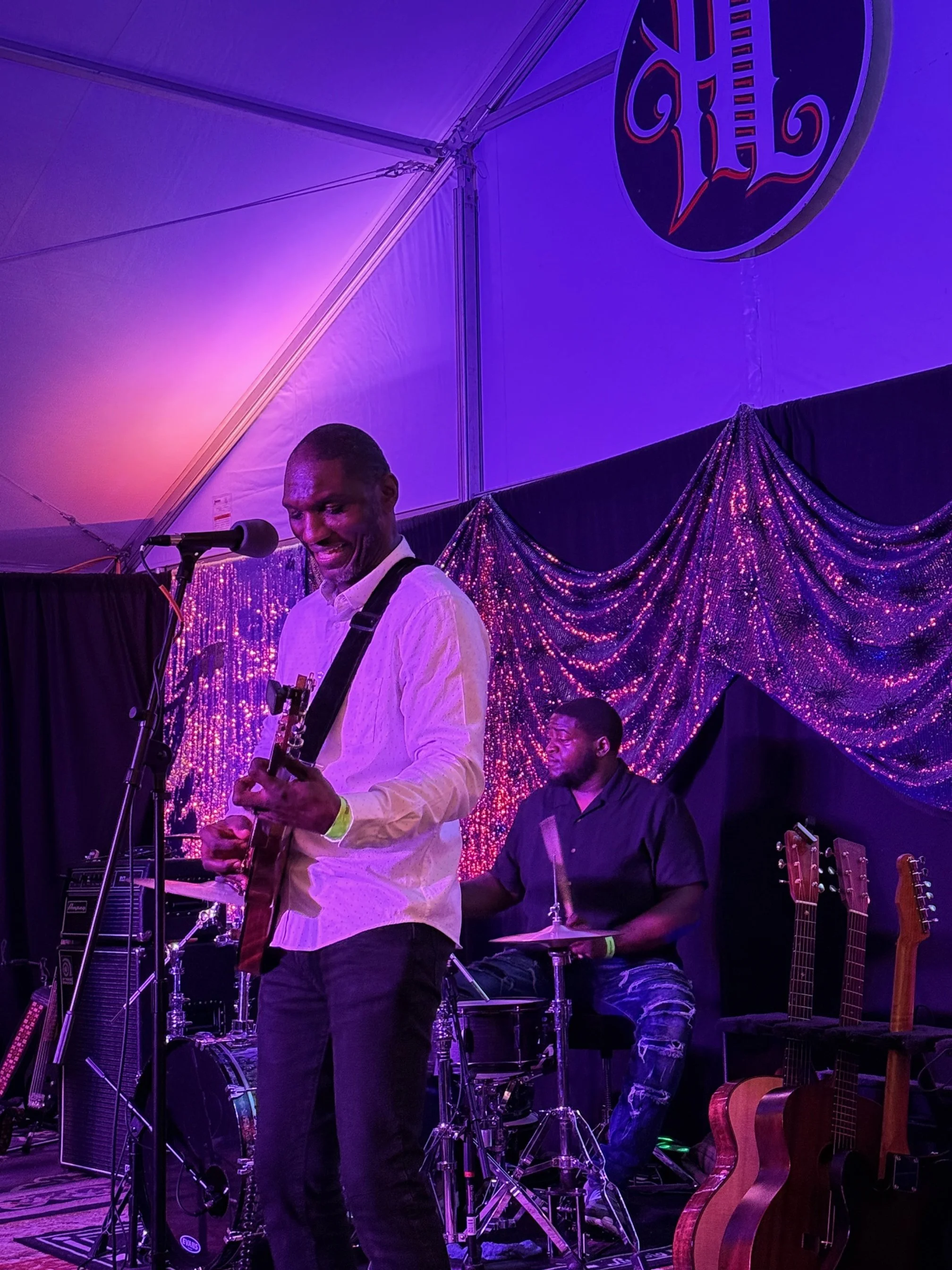 Musicians performing on stage under purple lighting, with one playing the guitar and the other on drums, surrounded by guitars and musical equipment.