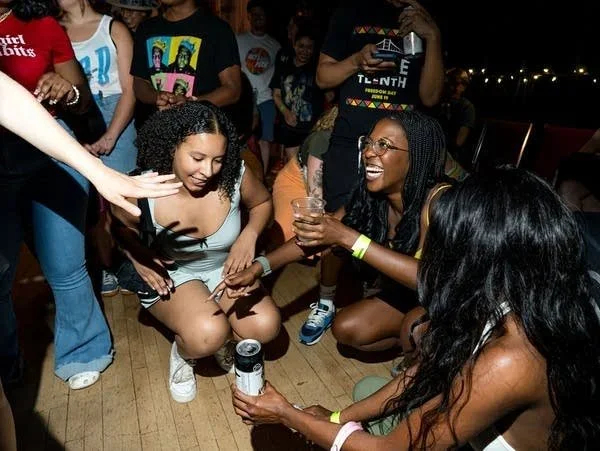 A group of young women and girls at a party, some holding drinks, laughing, and sitting on the floor. One woman is reaching out to touch a can on the floor.