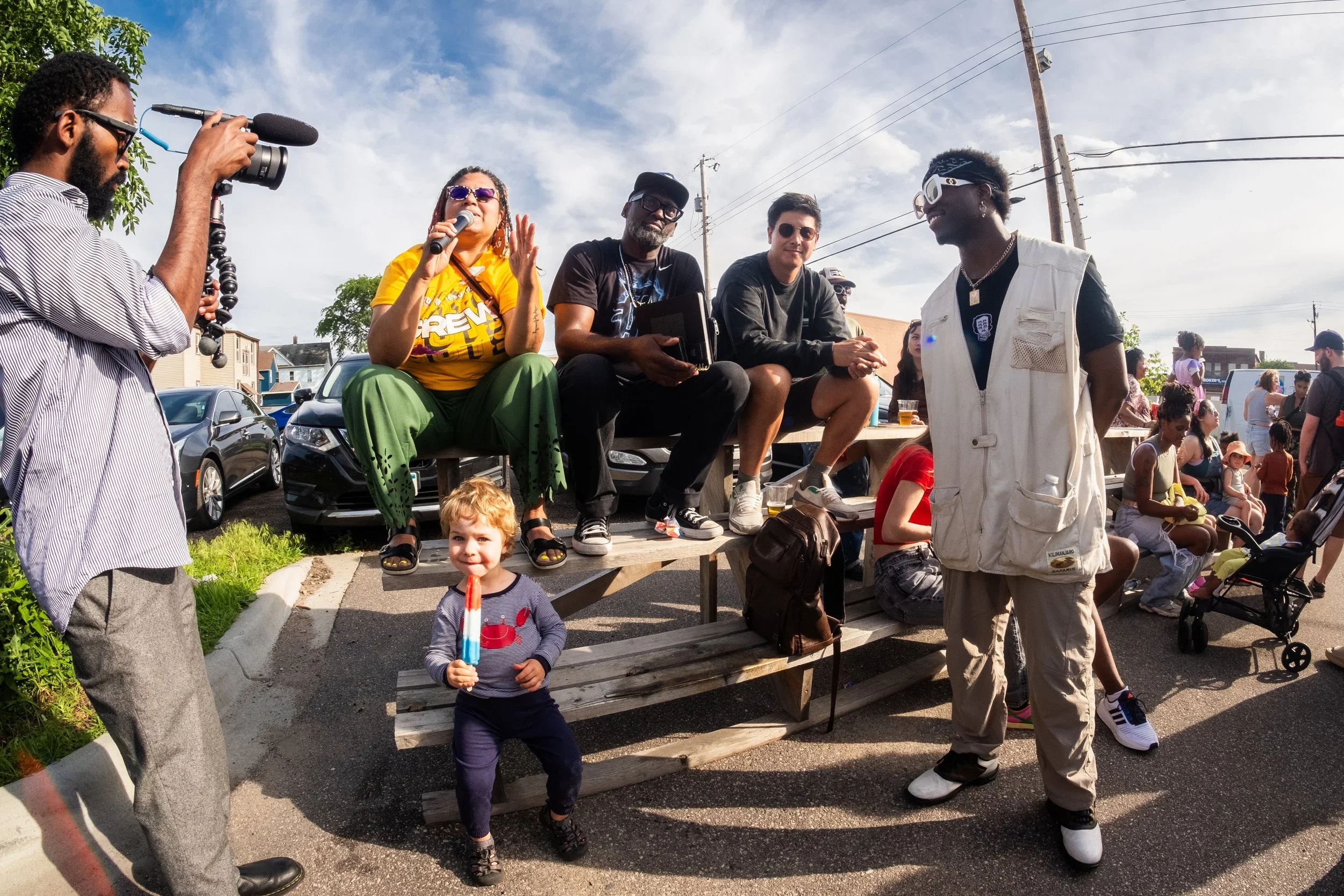 A group of people gathered outdoors during a community event, including a woman with sunglasses and a microphone speaking, people sitting on a wooden bench, a man with a camera recording, a smiling young child holding a popsicle, and other adults and children in the background.