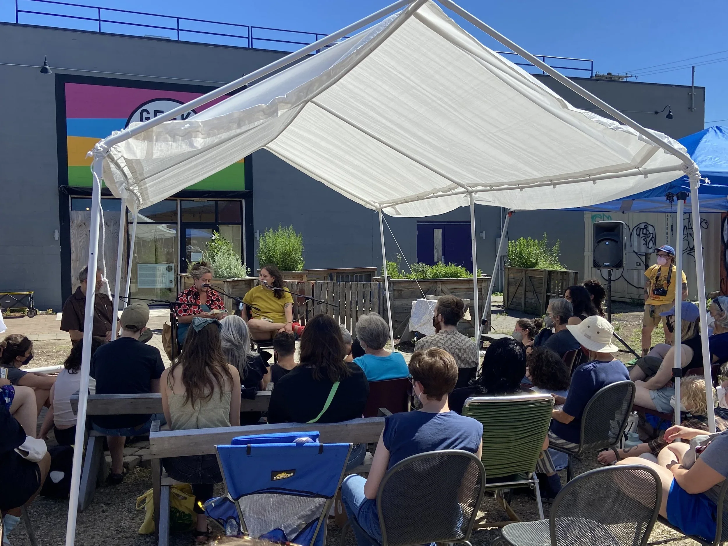 A group of people are gathered outdoors under a white tent, attending a panel discussion or interview with two women seated with microphones. The setting appears to be a community event or festival on a sunny day.