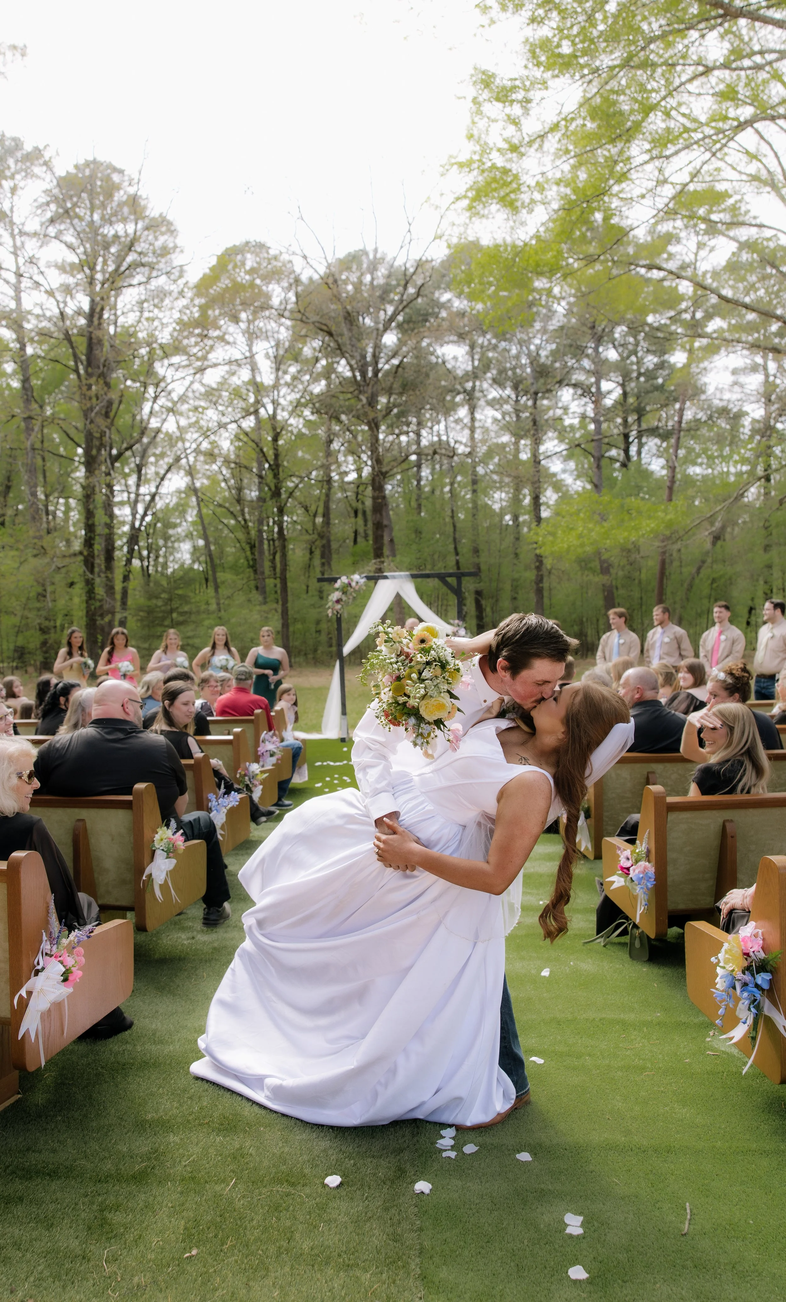 A newly married couple sharing a kiss outdoors during a wedding ceremony, surrounded by friends and family, with wedding decorations and trees in the background.