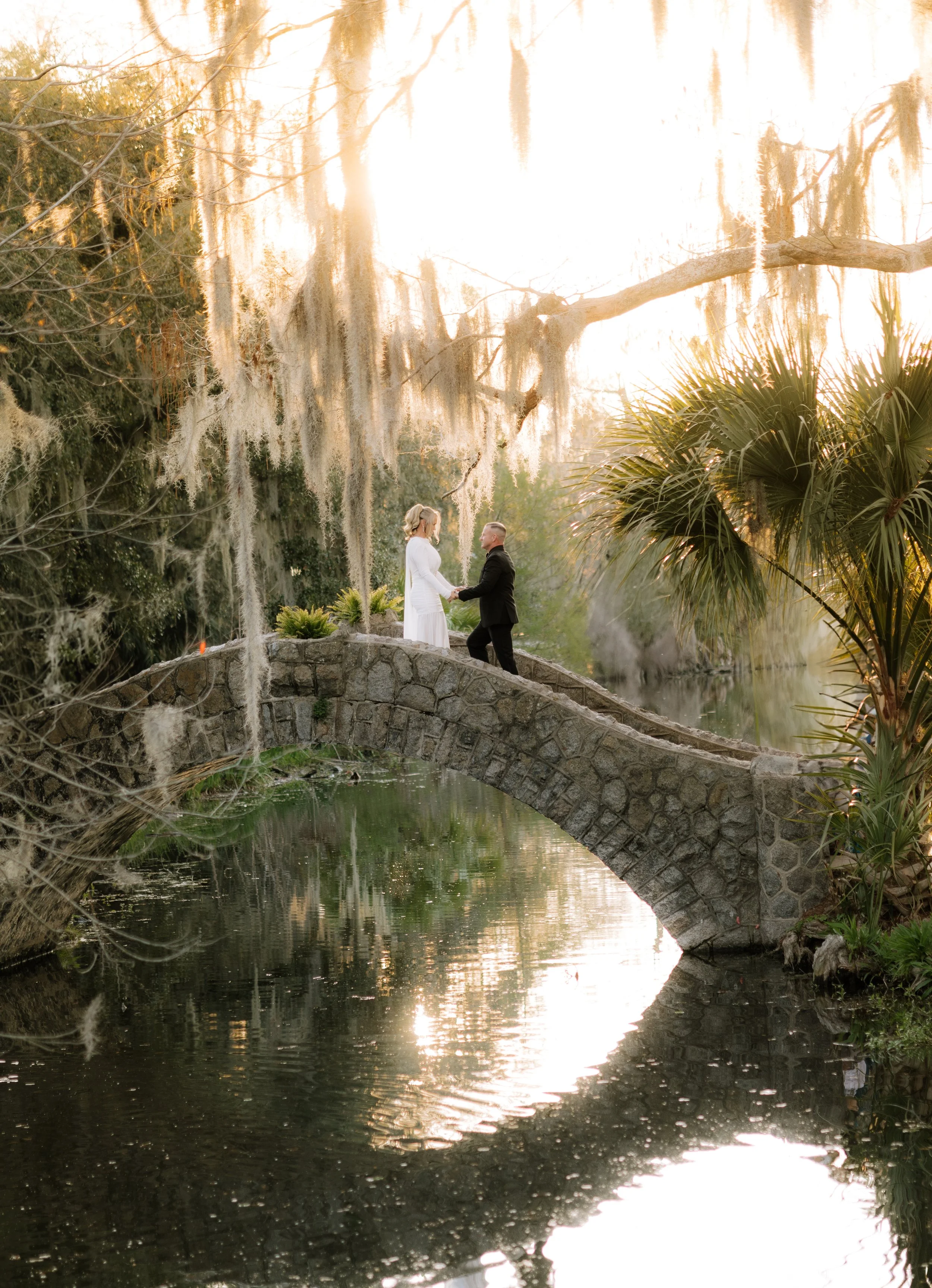 A couple standing on a stone bridge over a small pond, surrounded by lush trees with Spanish moss hanging from their branches, during sunset.