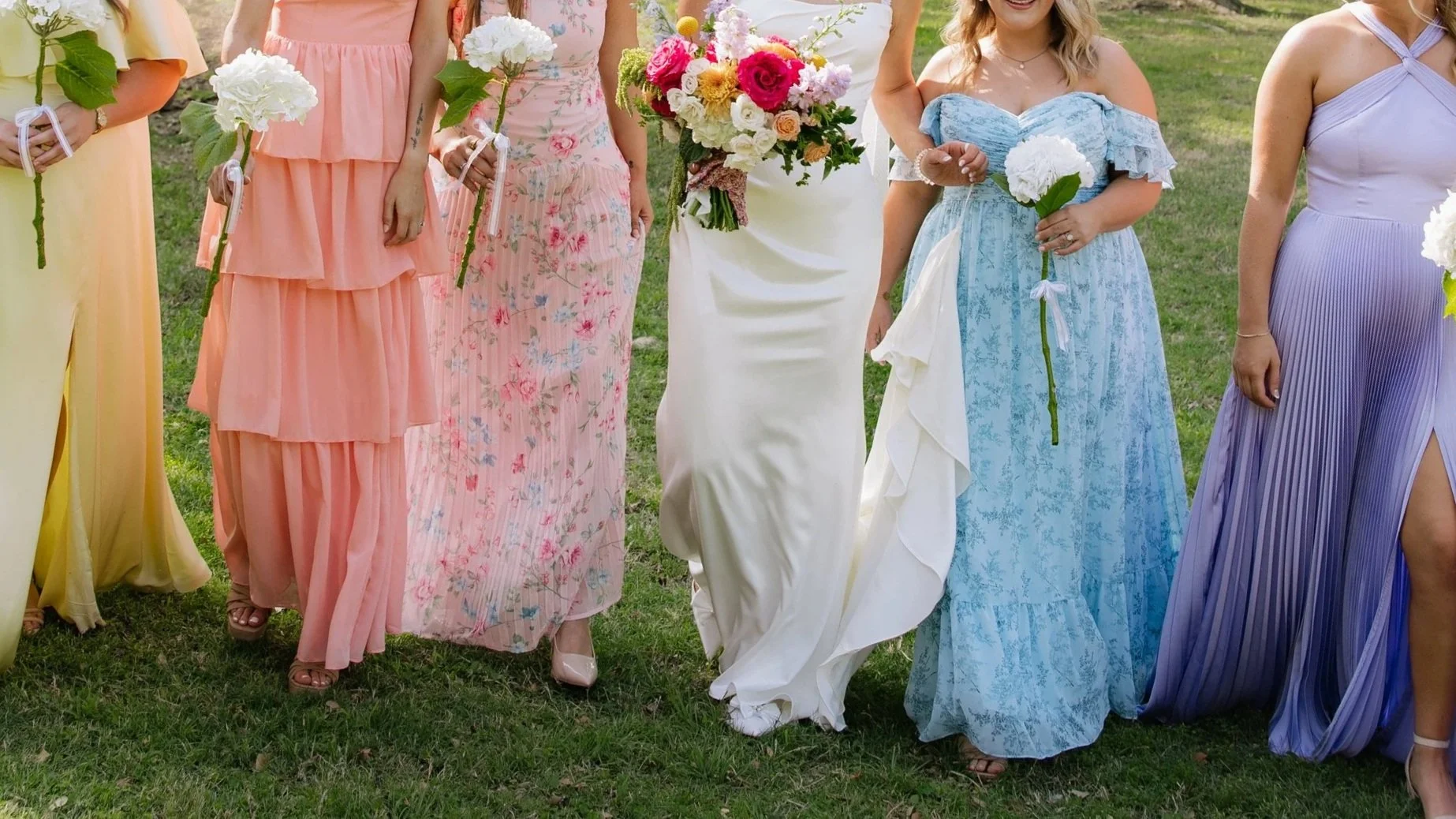 Women dressed in colorful floral dresses and holding bouquets of flowers, standing on grass outdoors, likely participating in a wedding or celebration.