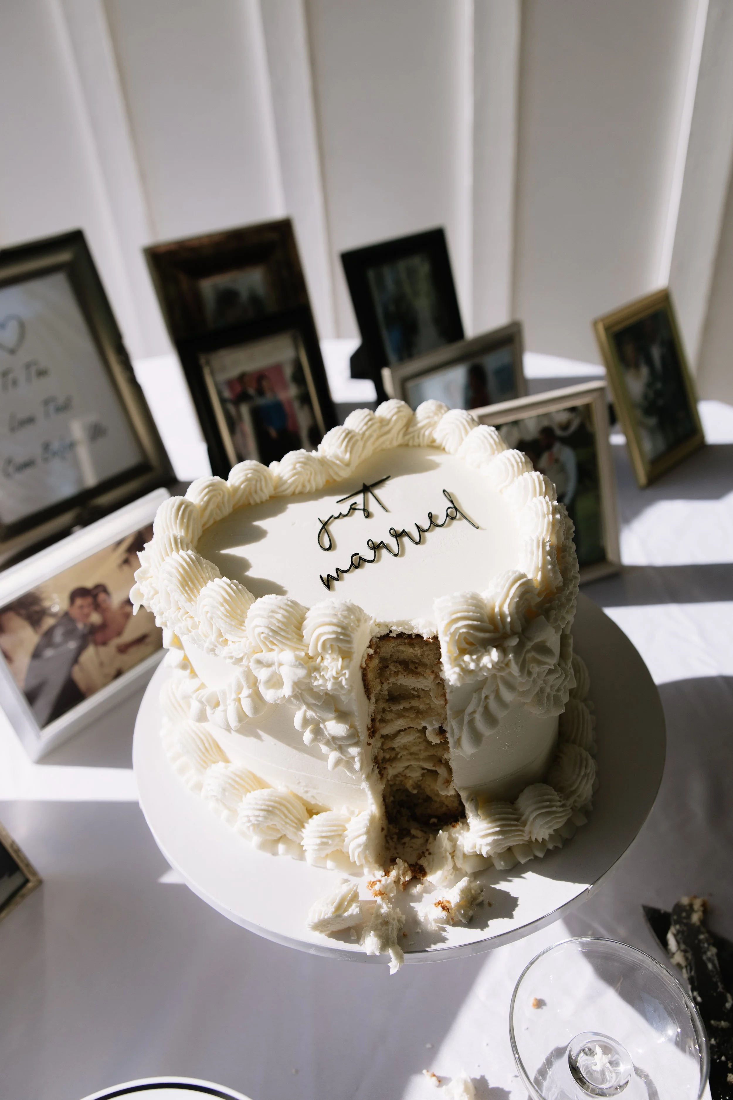 A wedding cake with a bite taken out, decorated with white frosting, sitting on a table with picture frames and empty glasses in the background, and a message that reads 'just married' on top.