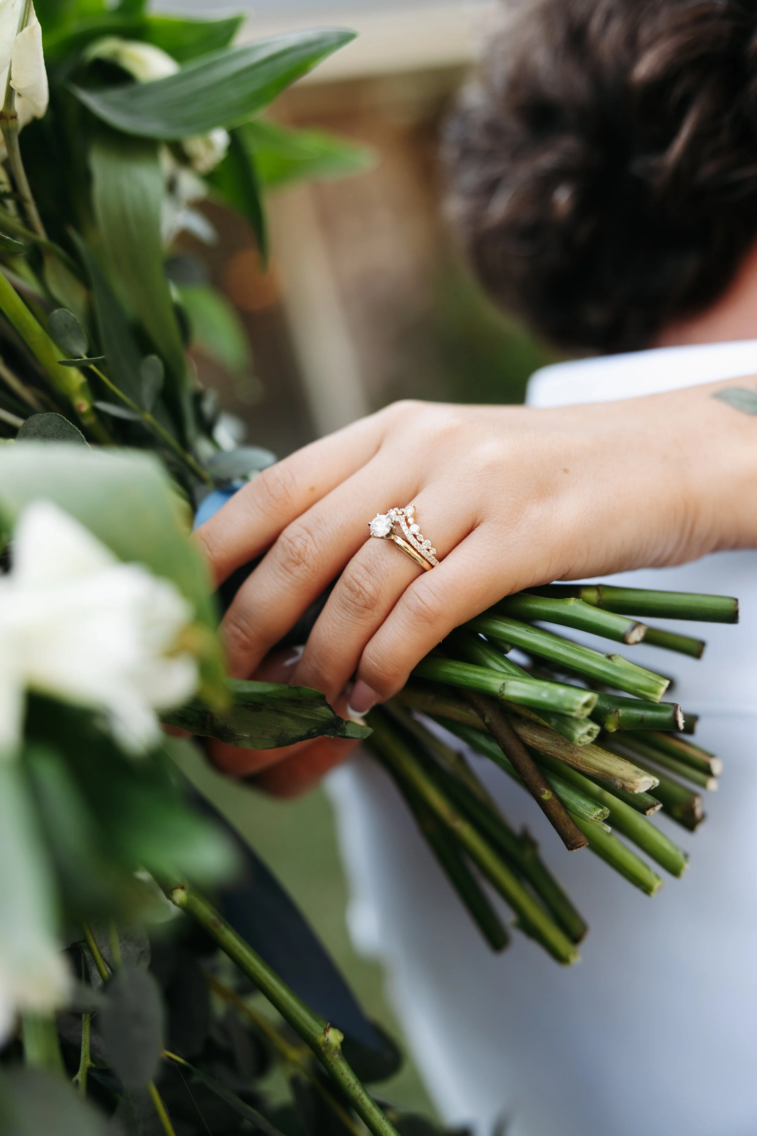 Close-up of a person's hand holding a bouquet of white flowers and greenery, with a focus on a wedding ring on the person's finger. The background is blurred.