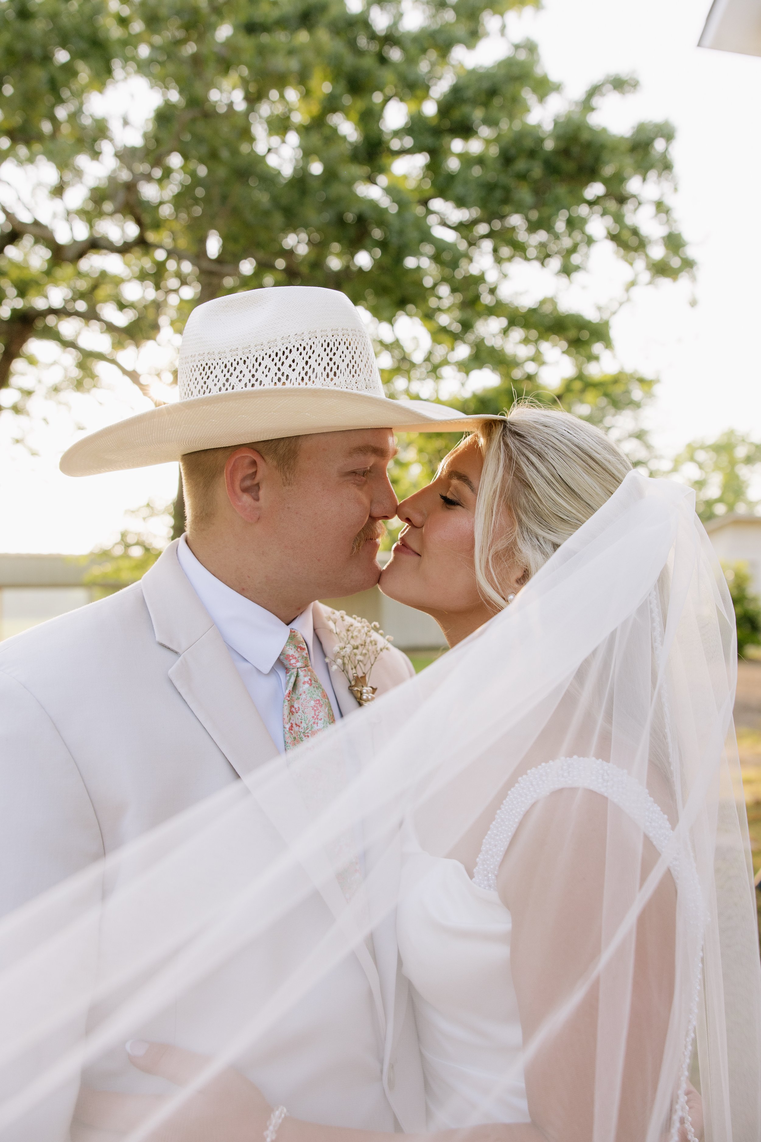 A bride and groom in wedding attire, about to kiss, outdoors with trees in the background, the groom wearing a cream-colored suit and a large white cowboy hat, and the bride in a white wedding dress with a veil.
