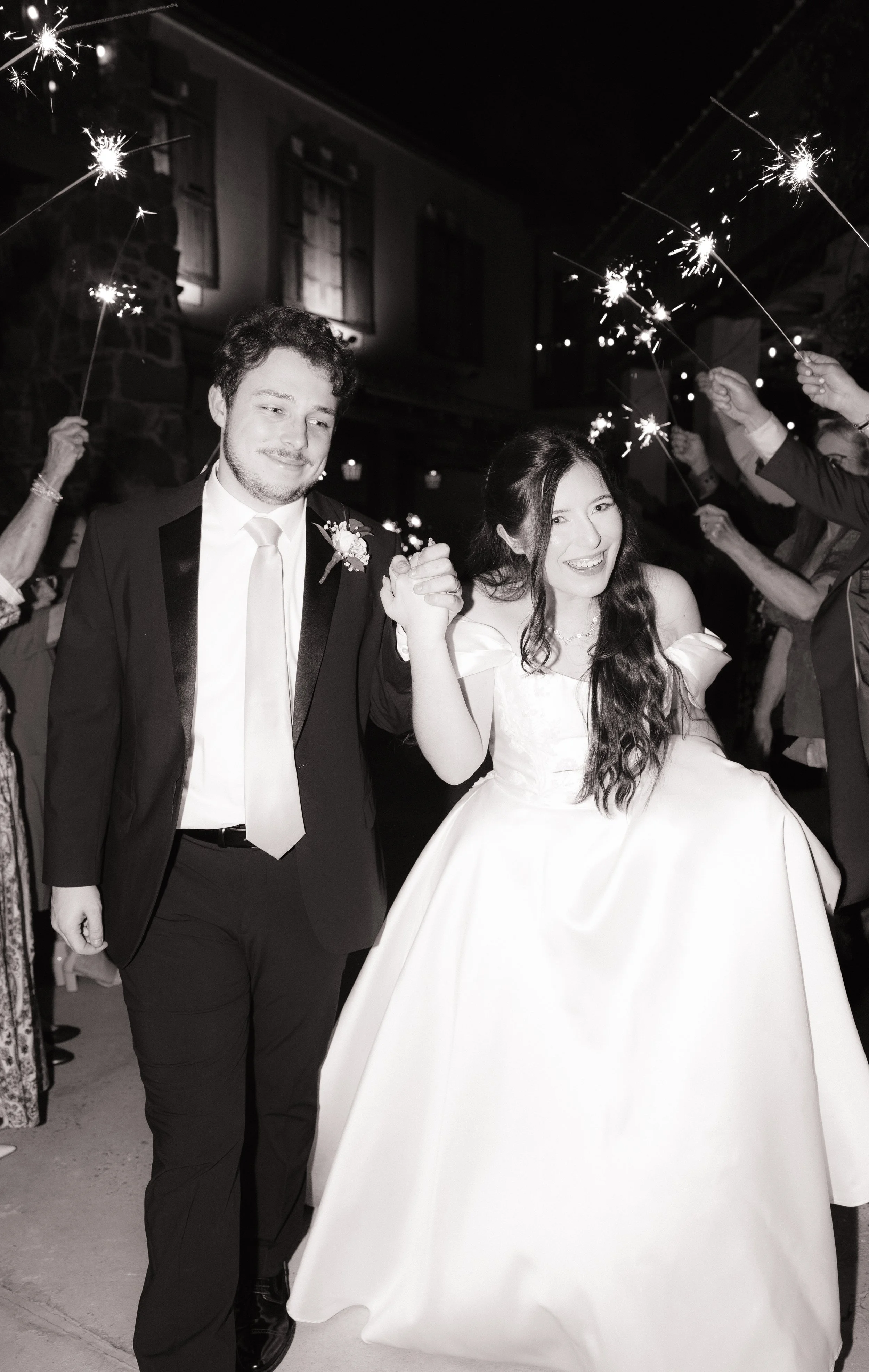 A black and white photo of a newlywed couple holding hands and walking at night. The groom is dressed in a tuxedo and the bride in a wedding gown. People around them are holding sparklers, celebrating outside a building with illuminated windows.