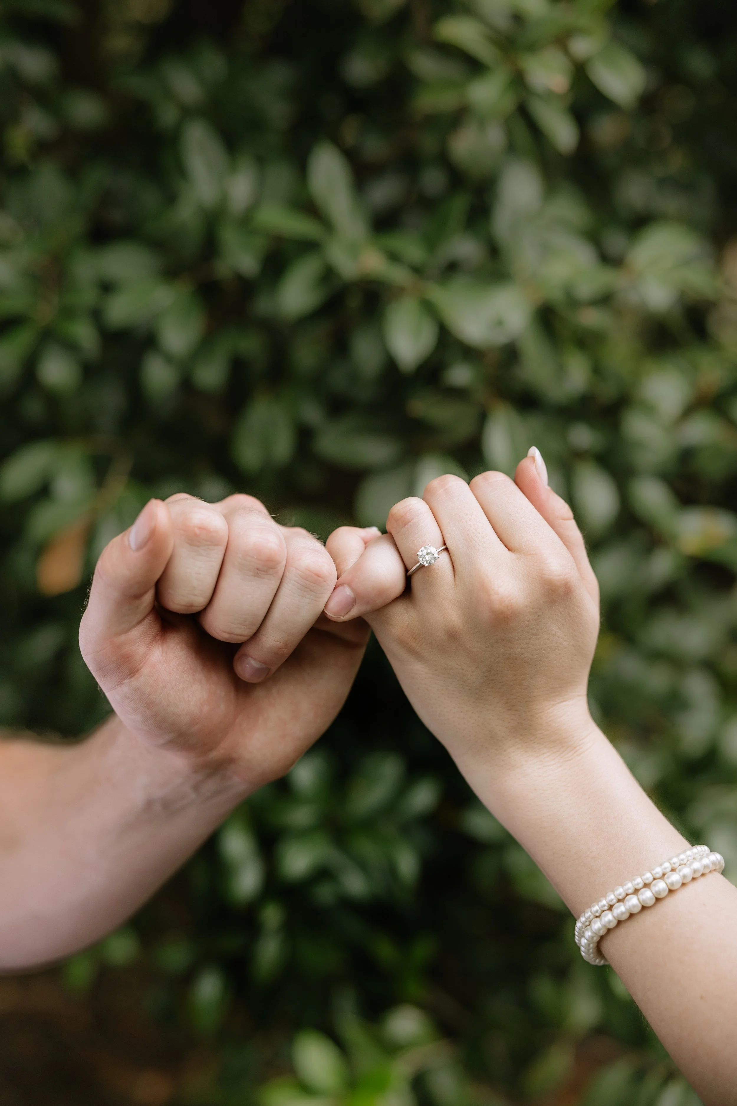Two hands, one with a diamond engagement ring, are held together in front of a green leafy background. The hand with the ring appears to be female, and the other hand appears to be male. The woman is also wearing pearl bracelets.