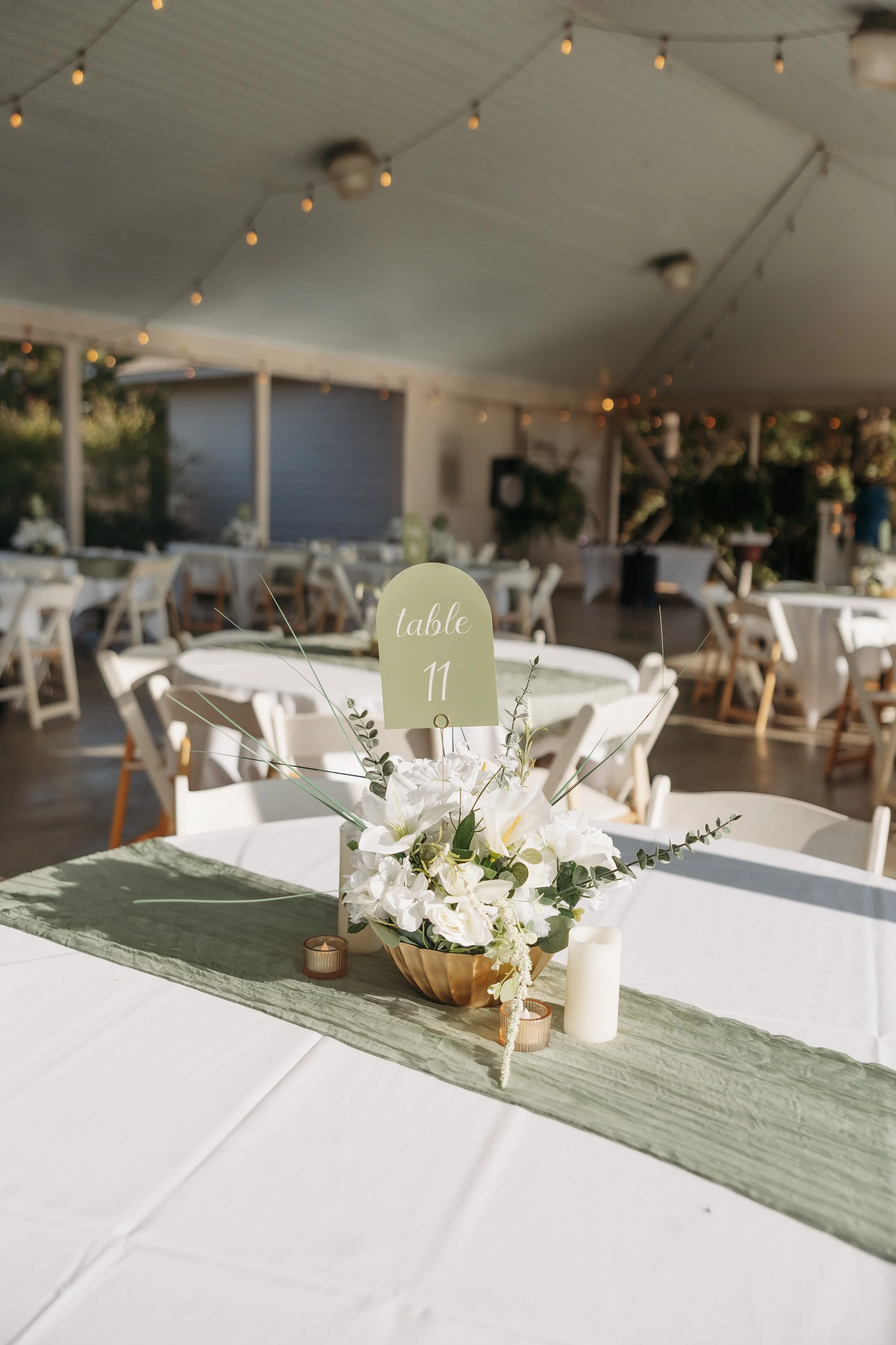 Wedding reception decorated with chairs, tables, and floral centerpieces including white flowers and greenery, with table number 11 sign, candles, and string lights overhead in a pavilion or outdoor venue.