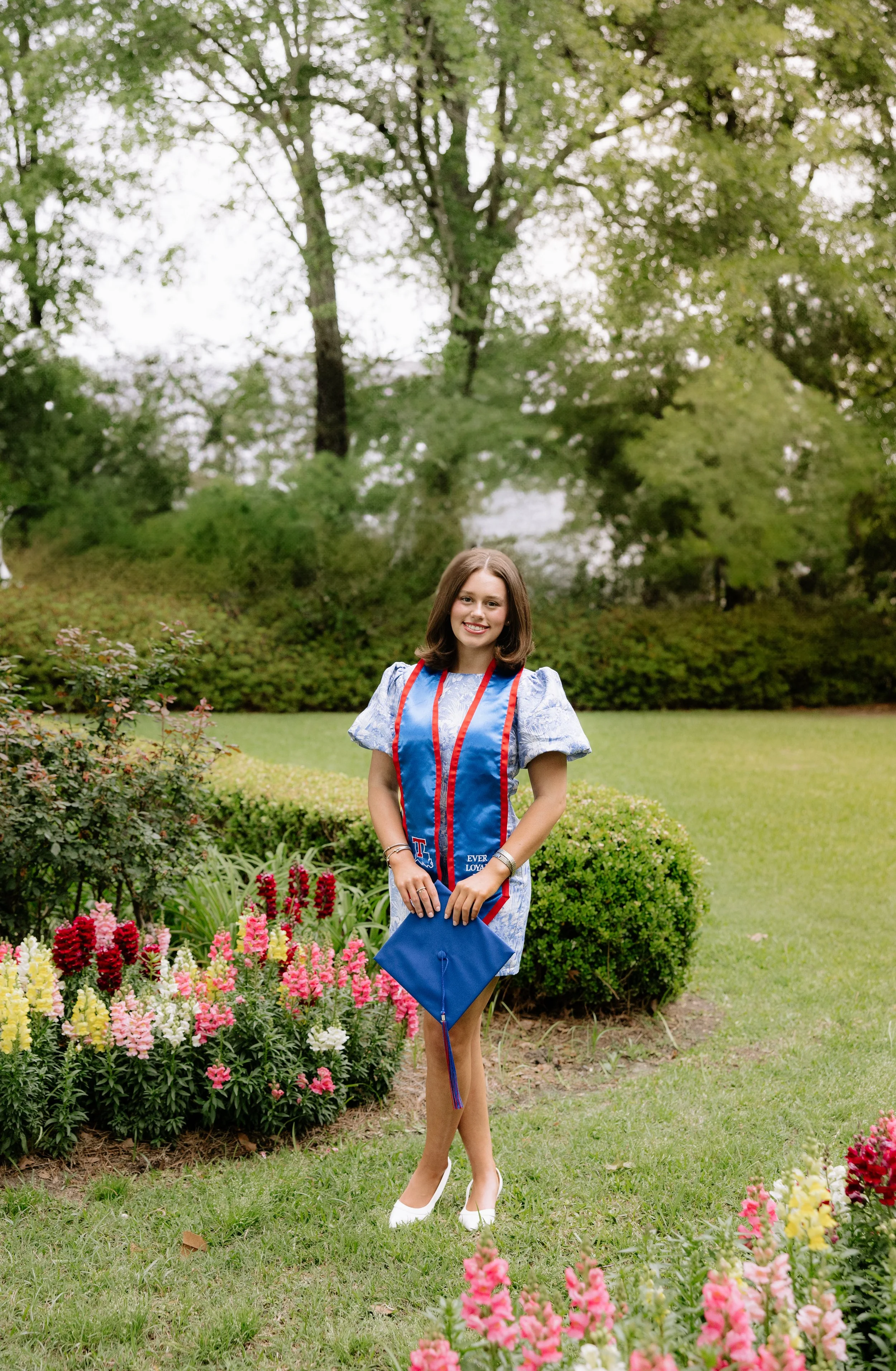 A young woman in a graduation gown and cap standing in a garden with colorful flowers and trees in the background.