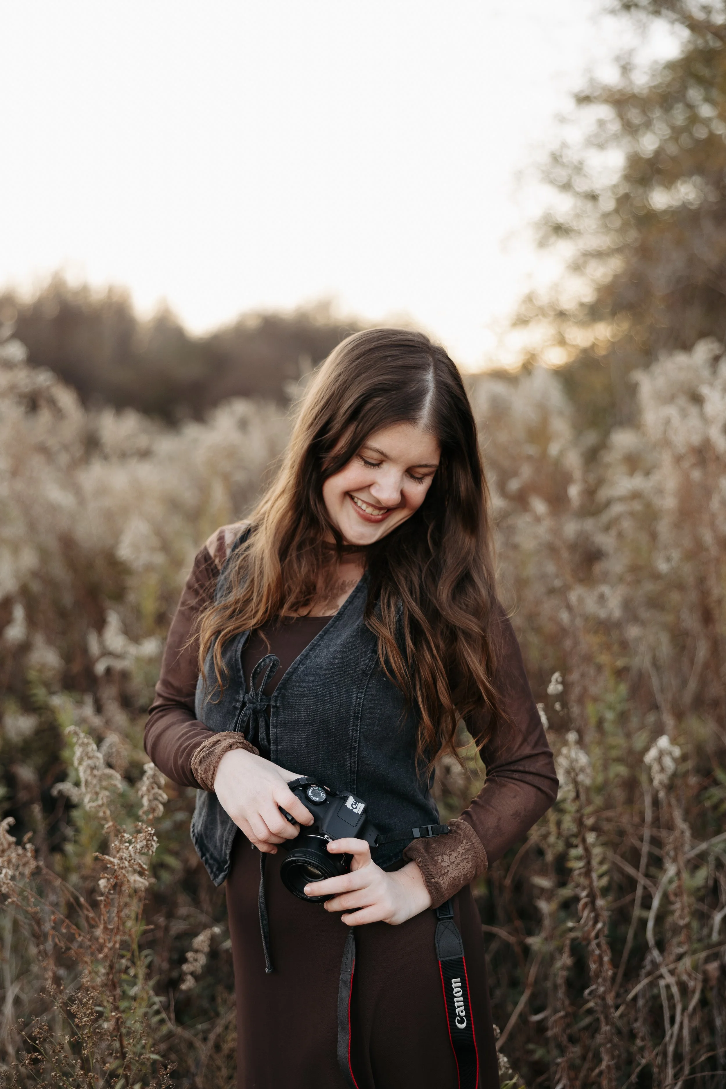 A woman with long brown hair, wearing a dark vest over a long-sleeve shirt, stands outdoors in a field of tall, dried plants, smiling while looking at a camera she is holding.