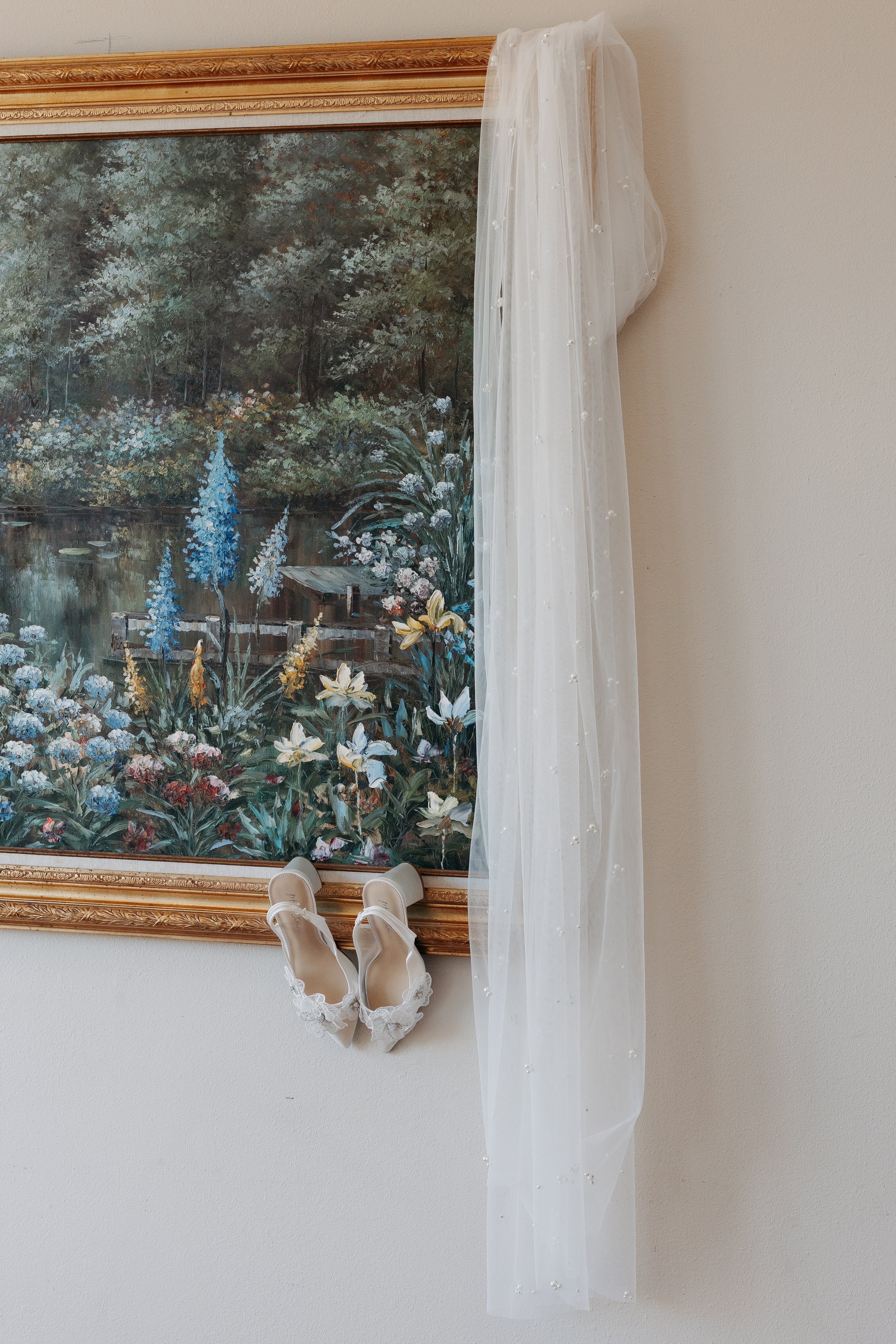 A framed landscape painting of a pond with trees and flowers hangs on a beige wall. A pair of white high-heeled shoes with lace details is attached to the bottom of the frame. Transparent fabric with small pearls is draped along the right side of the frame.