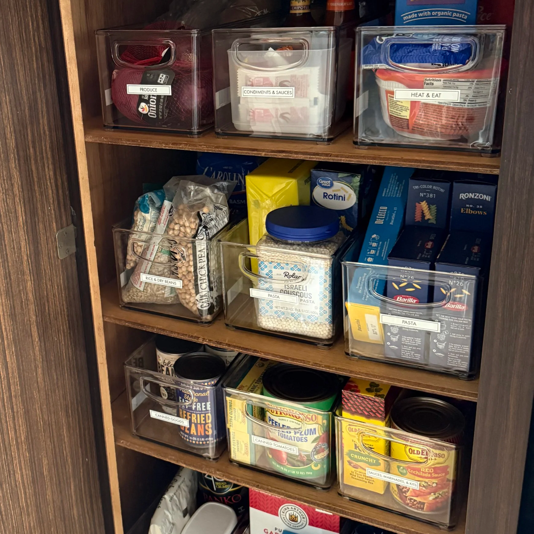 Open kitchen pantry with labeled clear bins containing produce, condiments, pasta, canned goods, and other food items.