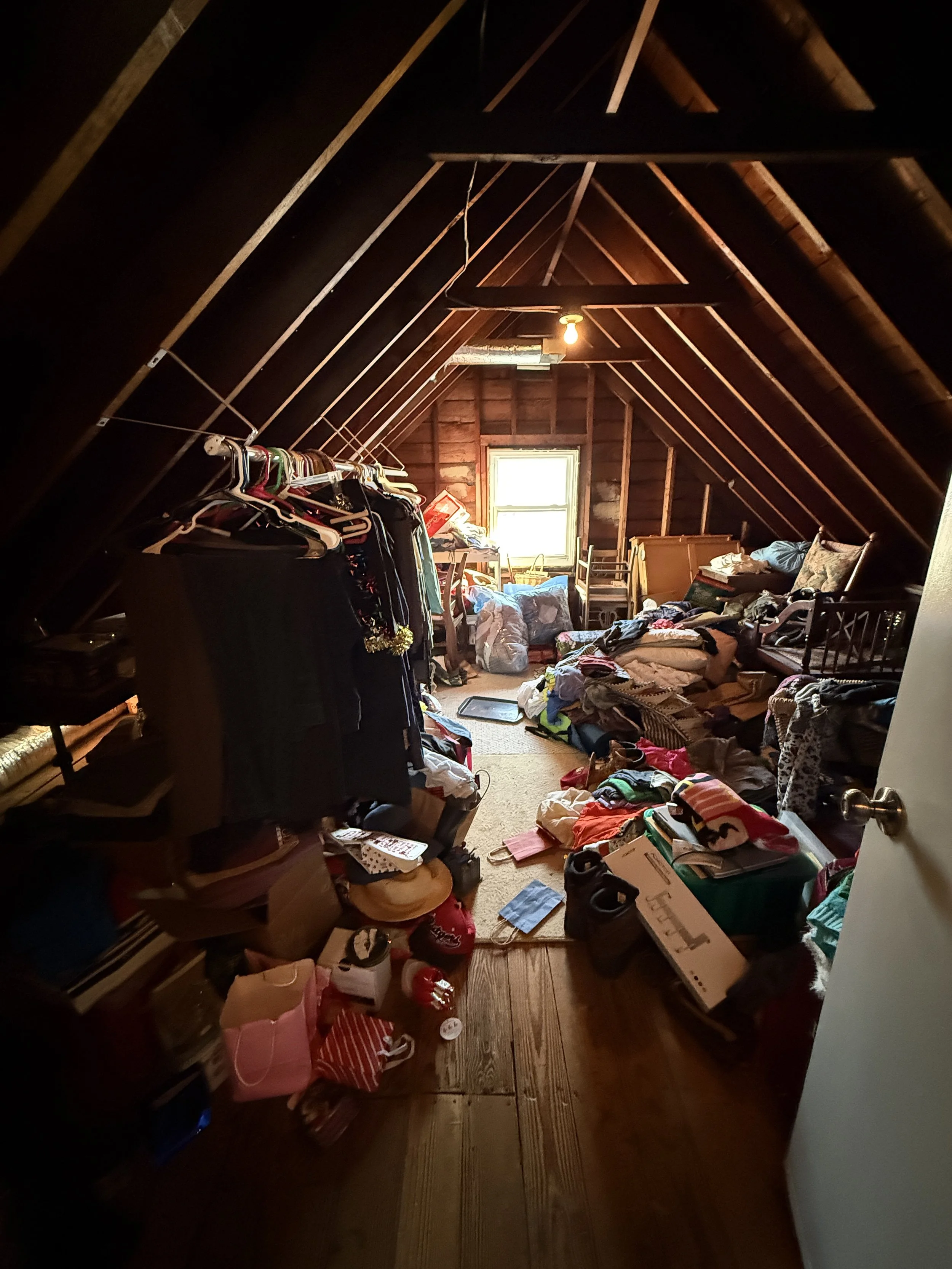 A cluttered attic with wood-paneled walls and a sloped ceiling, filled with clothes, boxes, and miscellaneous items, lit by a hanging light bulb and a small window at the back.