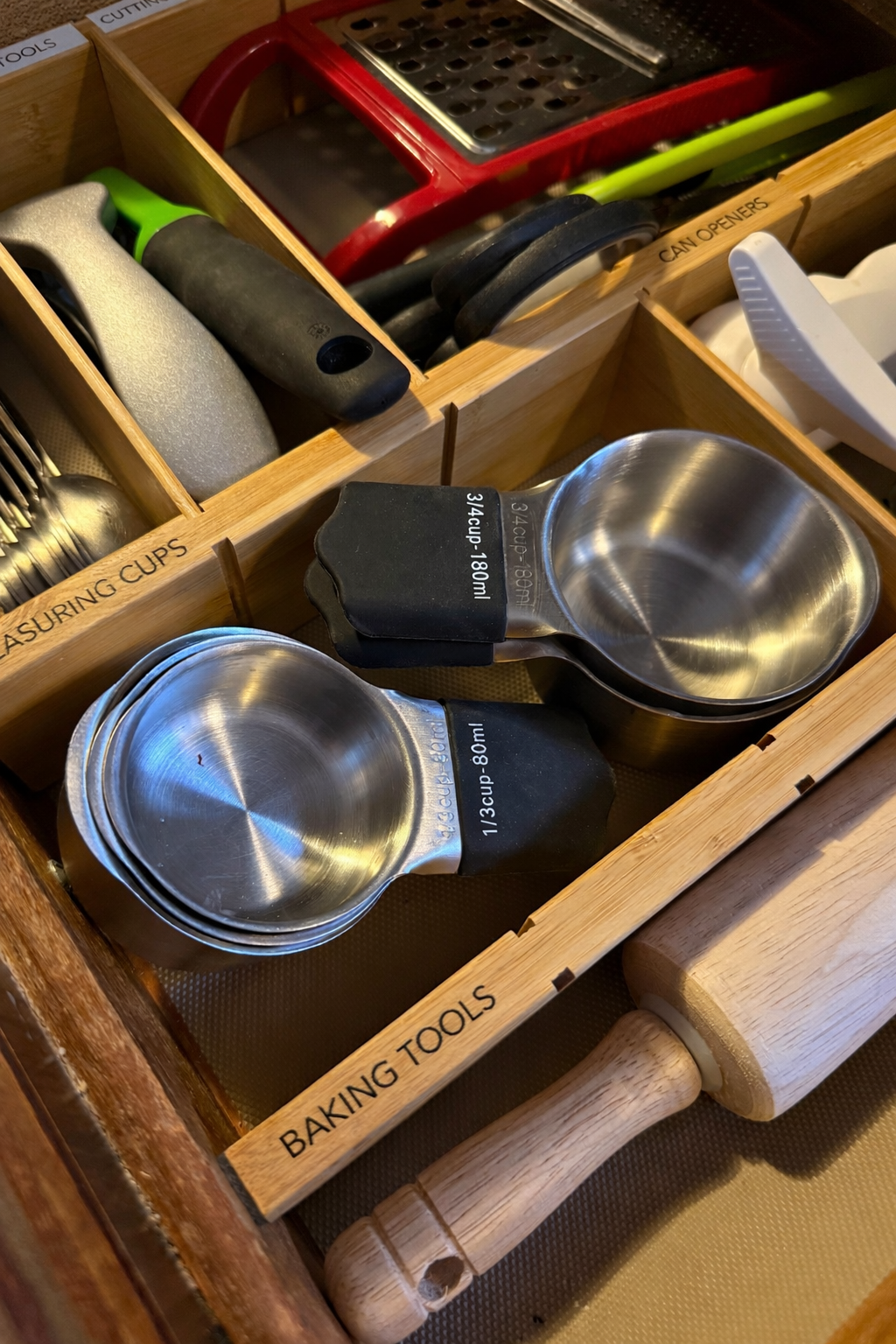 Wooden kitchen drawer divided into compartments labeled 'Measuring Cups,' 'Baking Tools,' and 'Can Openers.' Contains metal measuring cups, a meat tenderizer, and other kitchen utensils.