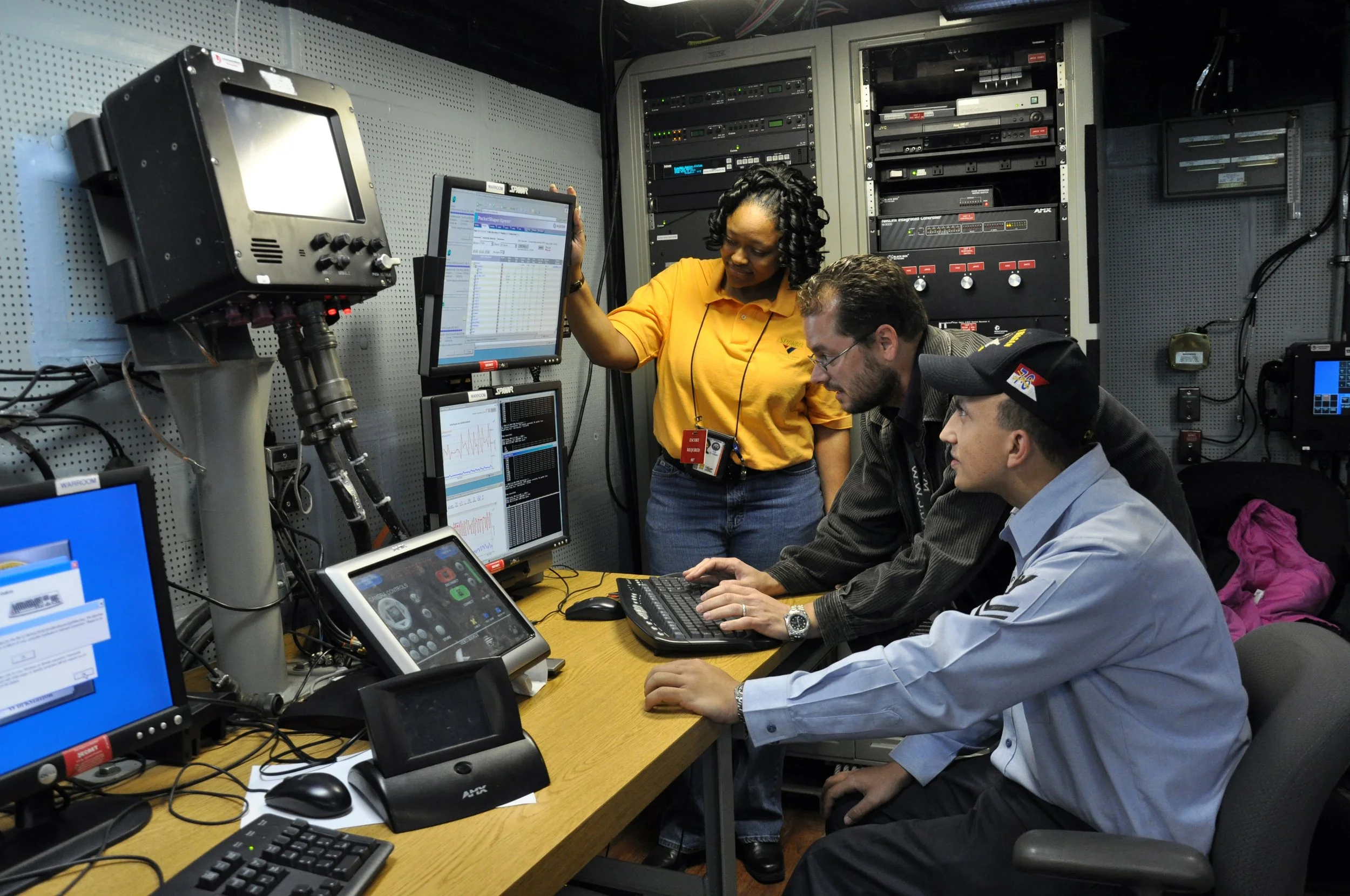 Three people working in a command center with multiple monitors and technical equipment. One woman stands pointing at a monitor, while two men sit at the desk working on computers.
