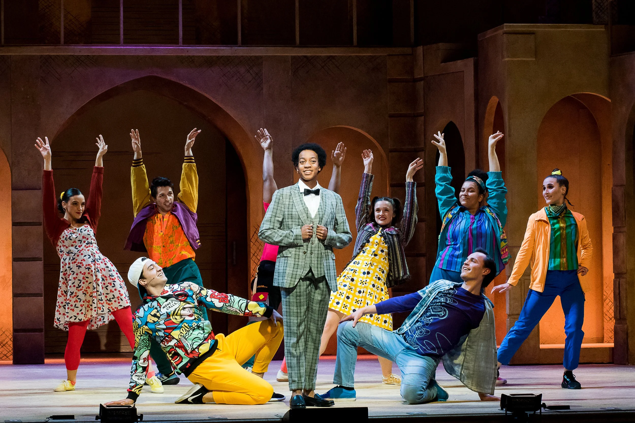A theatrical stage production featuring diverse performers in colorful costumes, with one person in a checkered suit and bow tie at center, surrounded by other actors in vibrant outfits, some standing with arms raised and others kneeling or sitting, 