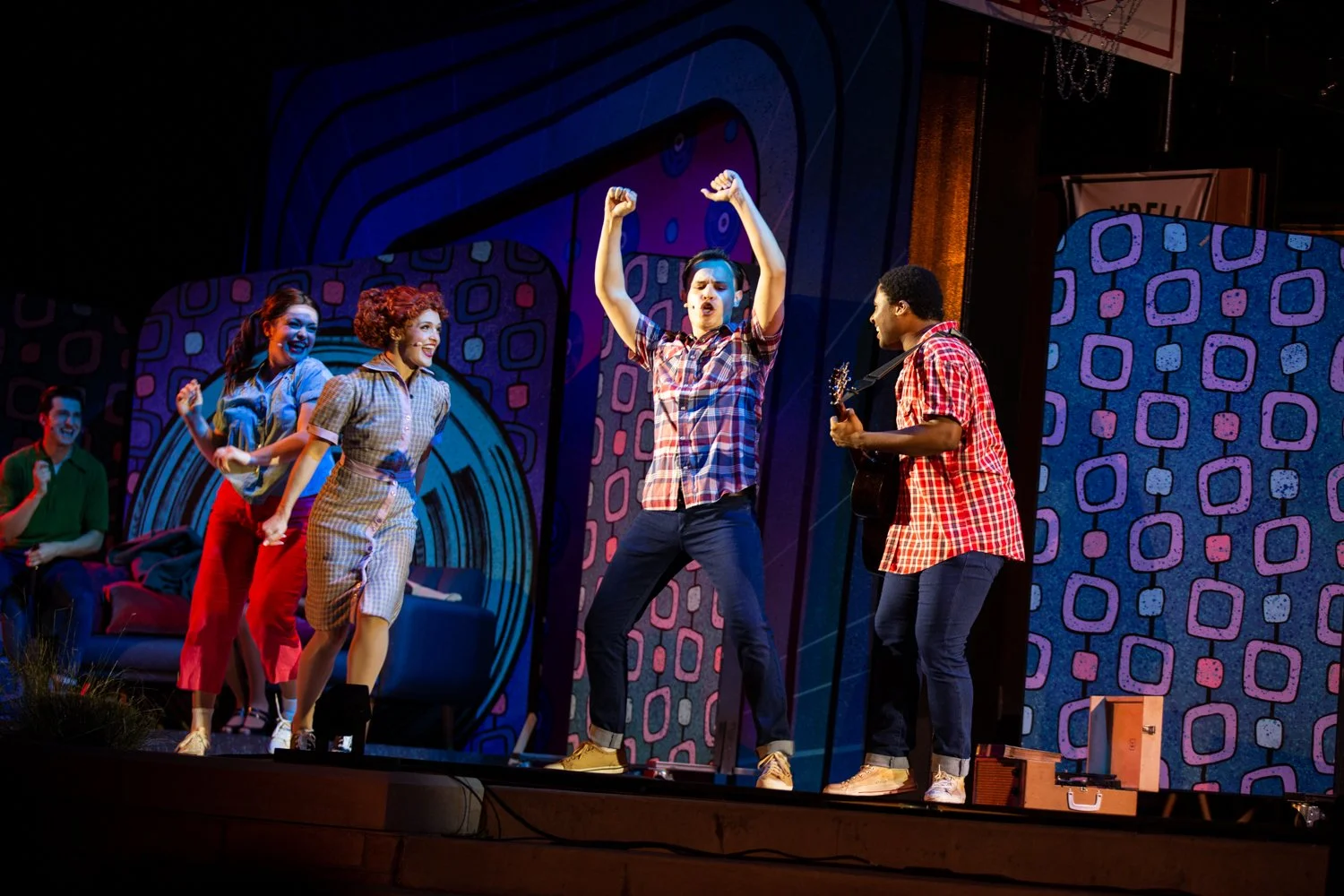 Theatre stage with five performers dressed in retro style, four women and one man playing guitar, dancing, and singing.