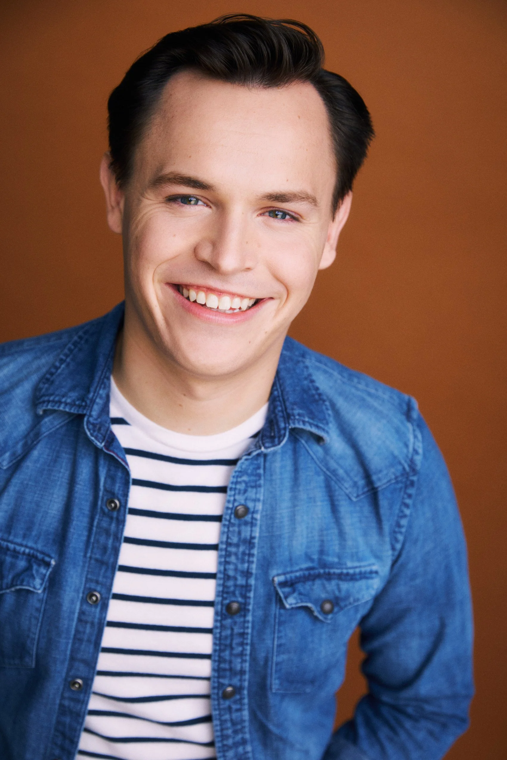Actor Connor Olney, smiling, wearing a blue denim jacket over a striped shirt, with a brown background.