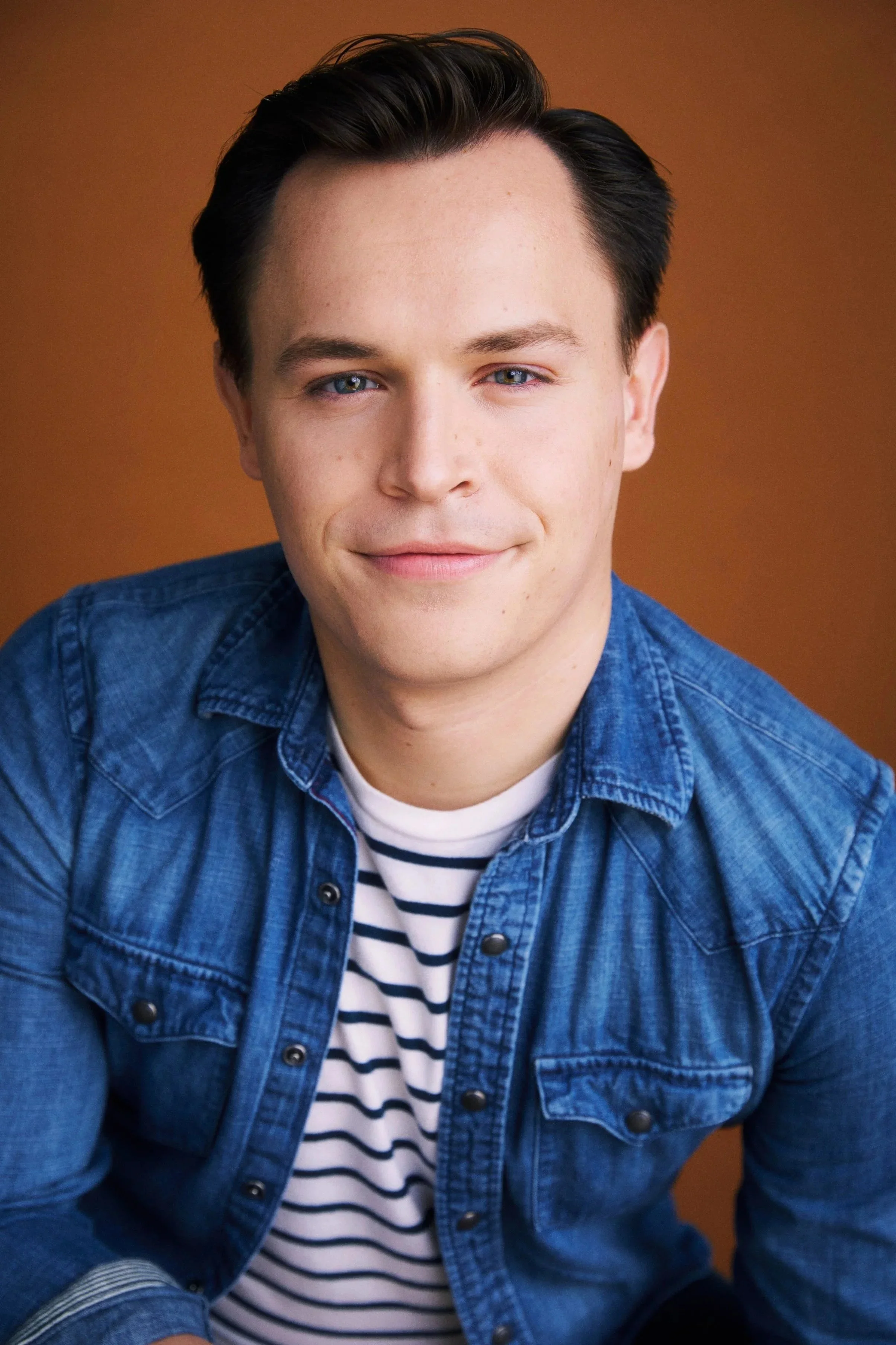 Actor Connor Olney, wearing a blue denim shirt over a striped white and navy shirt, against a warm brown background.