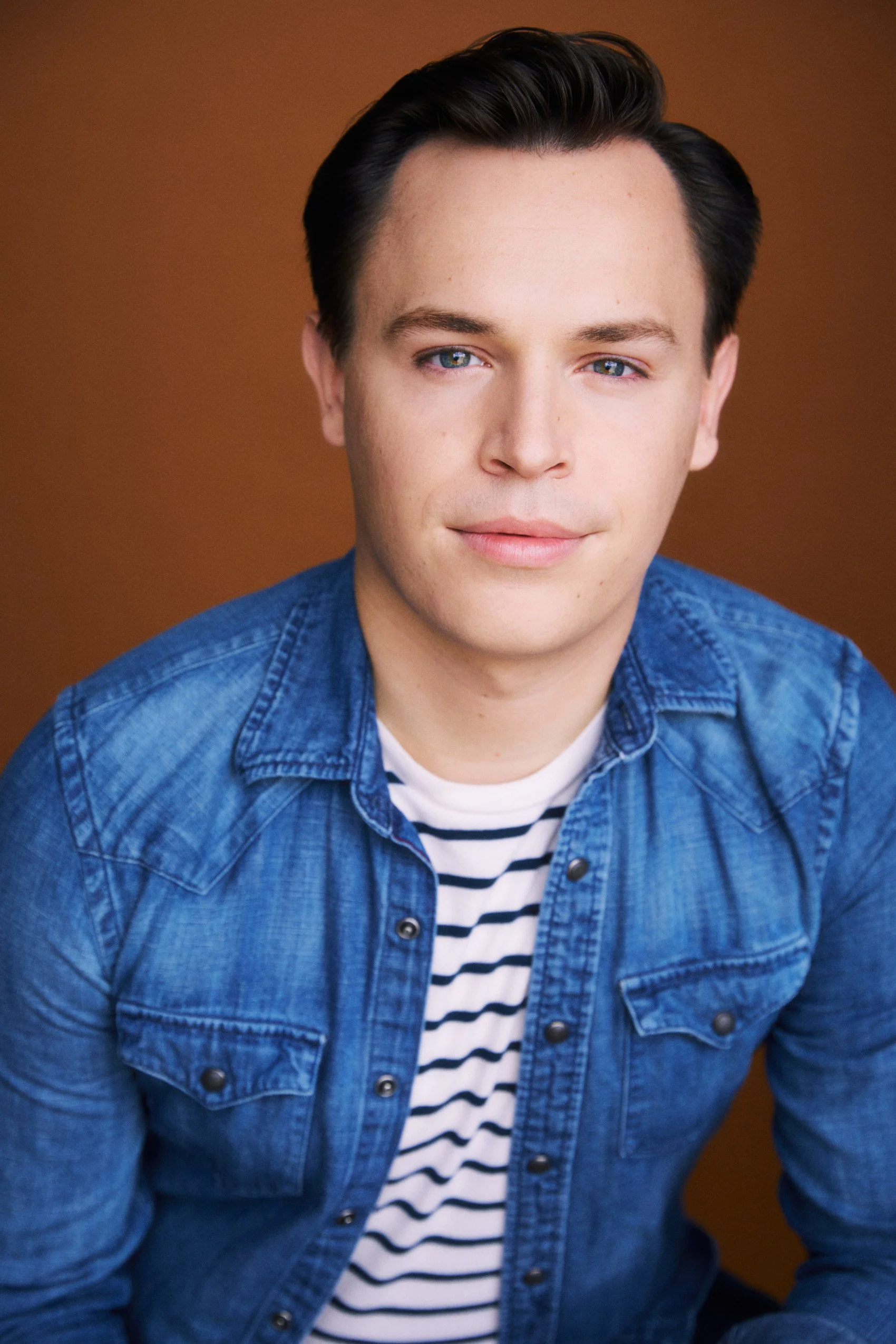 Actor Connor Olney, wearing a blue denim shirt over a white and navy striped t-shirt, posing against a brown background.