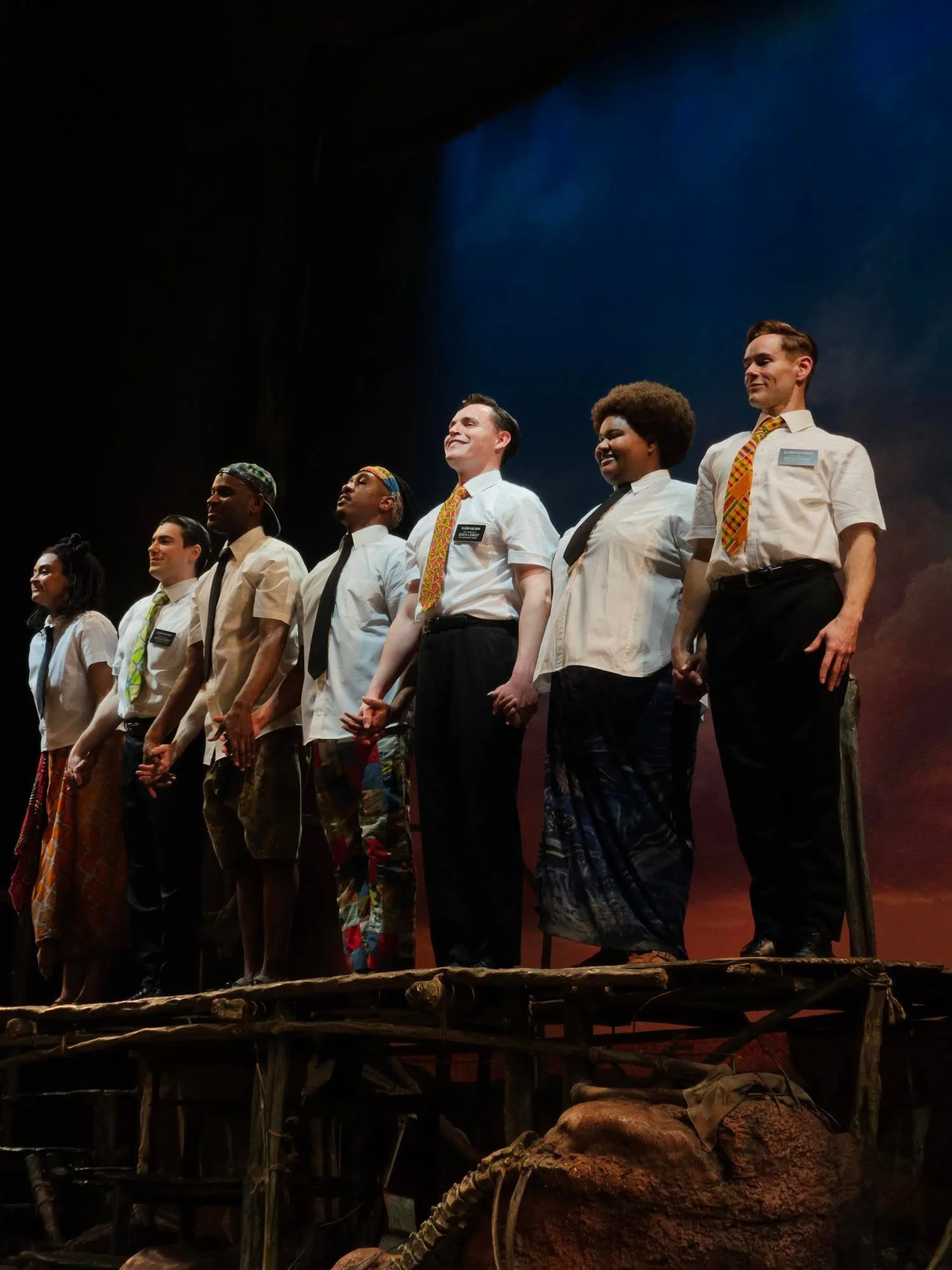 The Broadway cast of The Book of Mormon standing hand-in-hand on a stage with a colorful background, smiling and looking towards the audience.