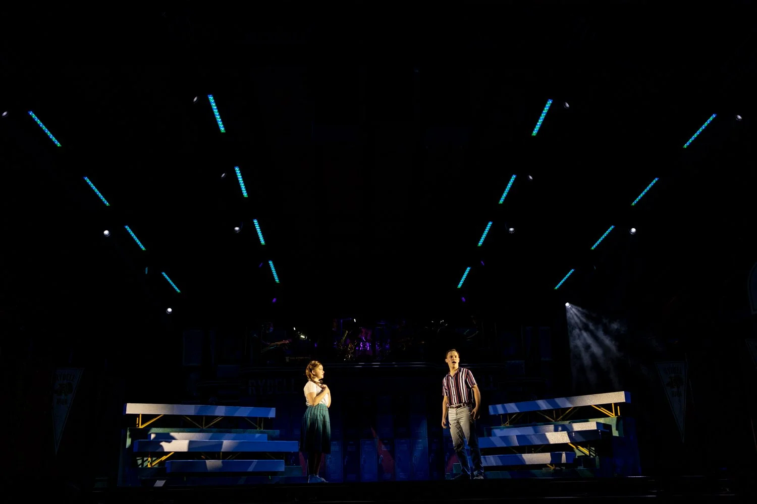 Two actors on stage in a theatrical production, with one woman and one man, surrounded by stage set pieces and painted backdrops.