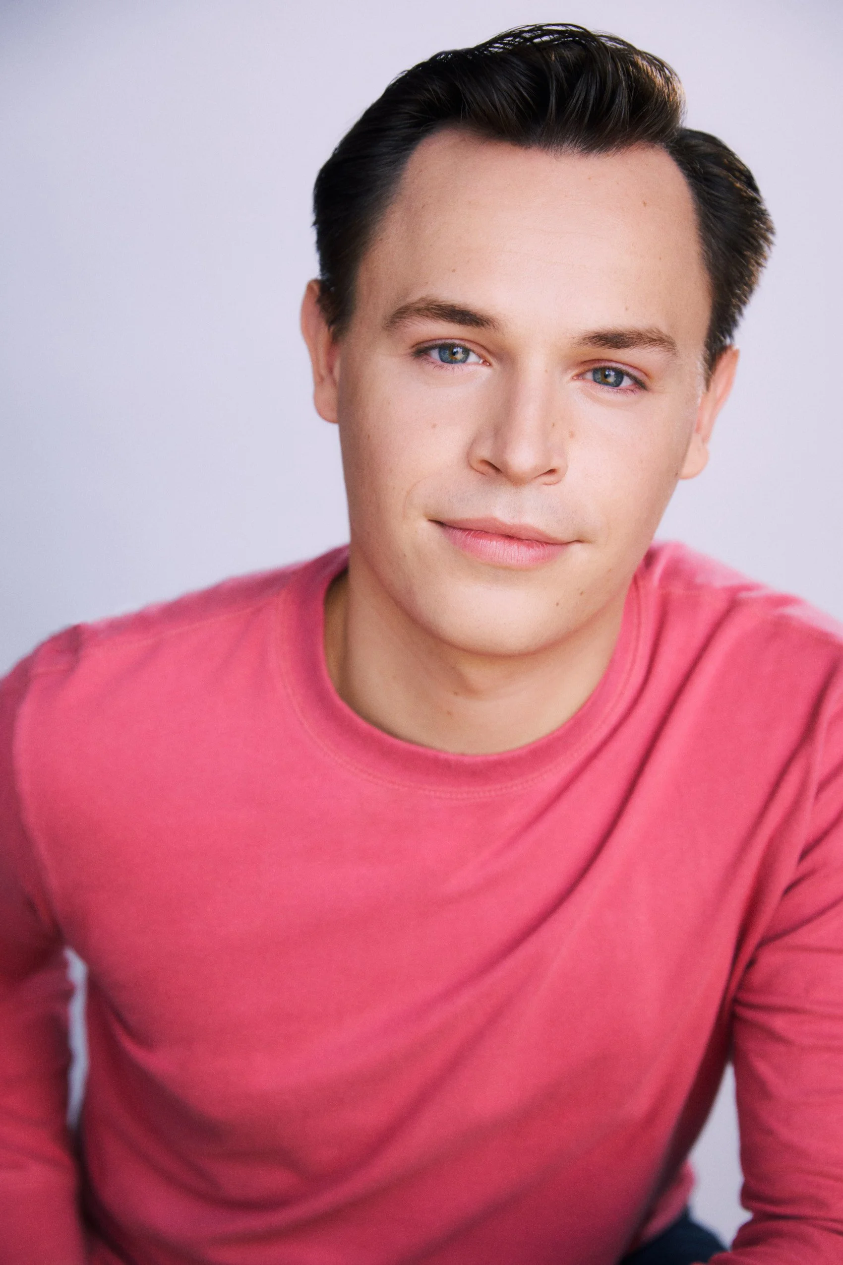 Actor Connor Olney, with short dark hair and blue eyes wearing a pink shirt, smiling softly, against a plain light background.