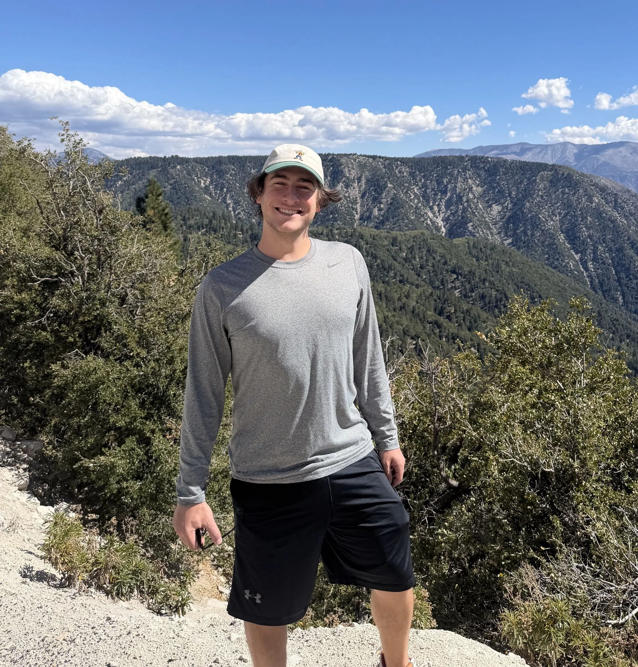 Young man smiling outdoors on a mountain trail, wearing a gray long-sleeve athletic shirt, black shorts, a baseball cap, and holding sunglasses, with mountains and partly cloudy sky in the background.